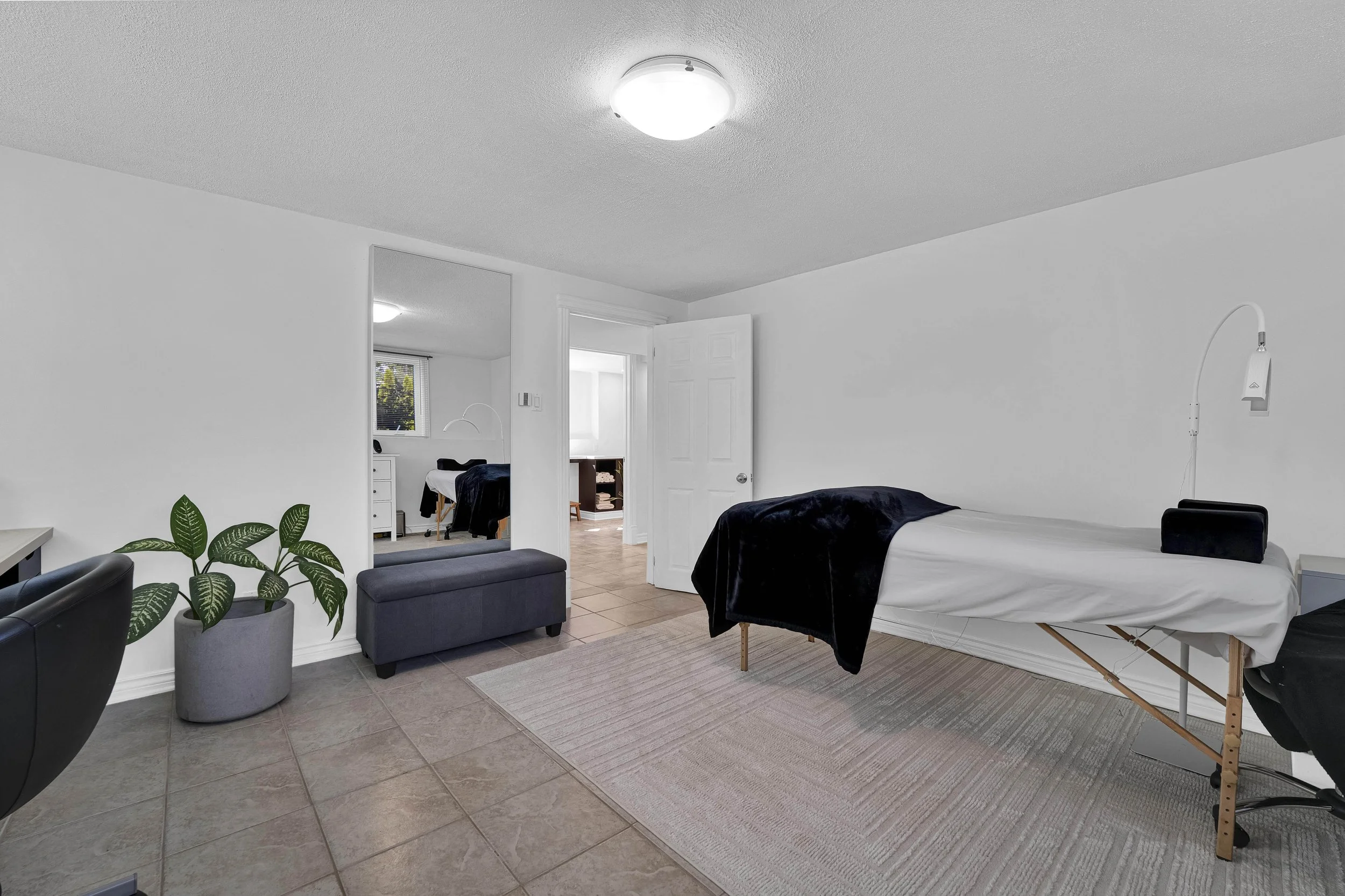 Ottawa Real EstateA bedroom with a bed, black and white bedding, a bedside table, a white cloth over the bed, a plant on the floor, and a rug, with an open door leading to another room with a desk and window, all with white walls and a ceiling light.