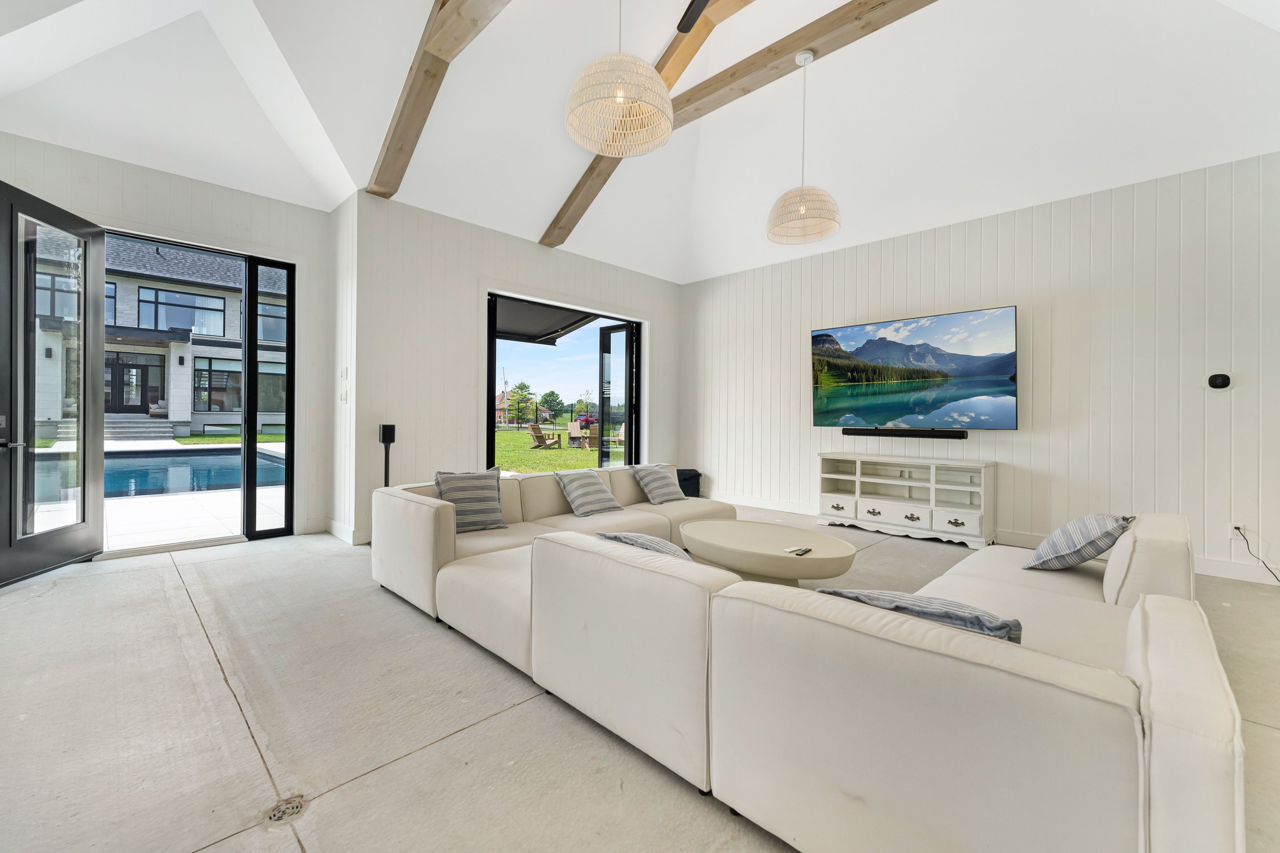 Modern living room with cream sectional sofa, wall-mounted TV displaying mountain scenery, open patio doors leading to a pool and outdoor area, wooden ceiling beams, and pendant lights.