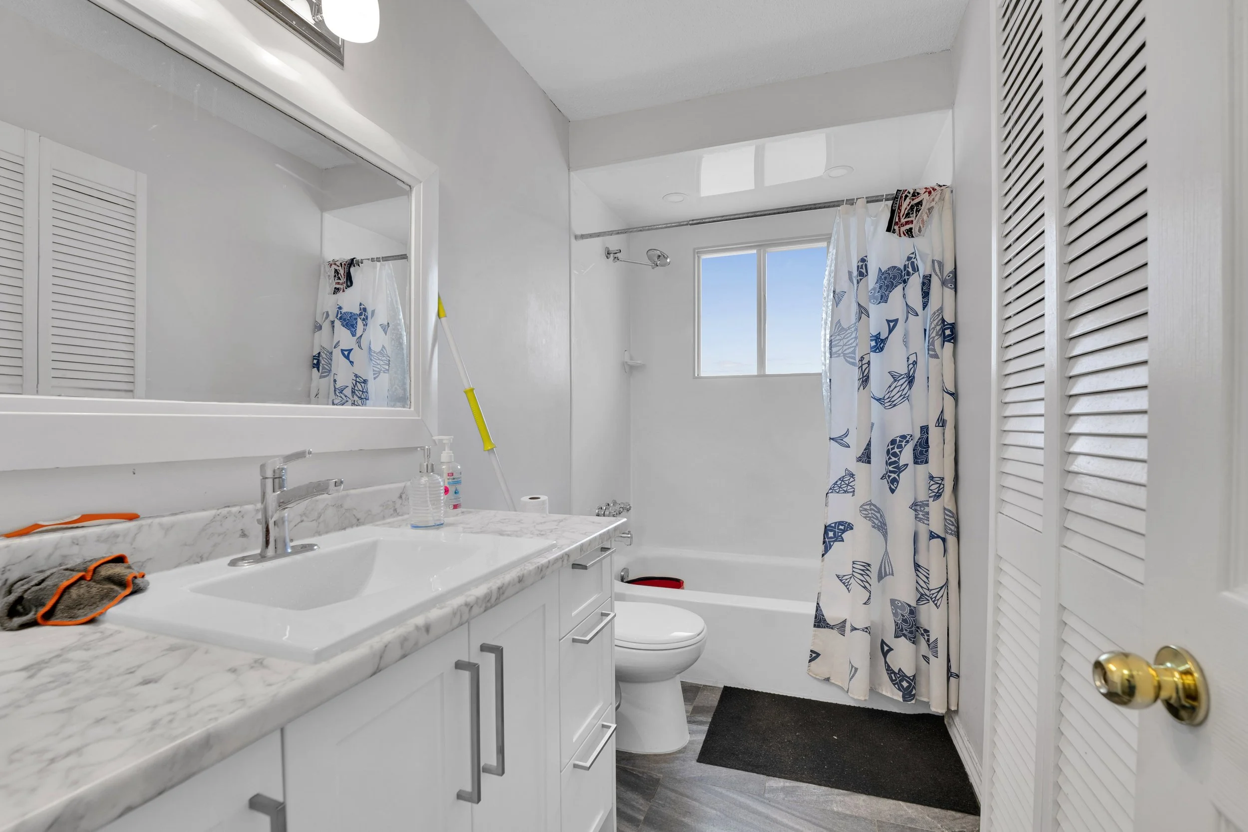 Ottawa Real Estate Media Services  A bathroom with a marble countertop sink, a mirror, a window showing a blue sky, a shower with a patterned curtain, a toilet, a black rug, and cleaning supplies.