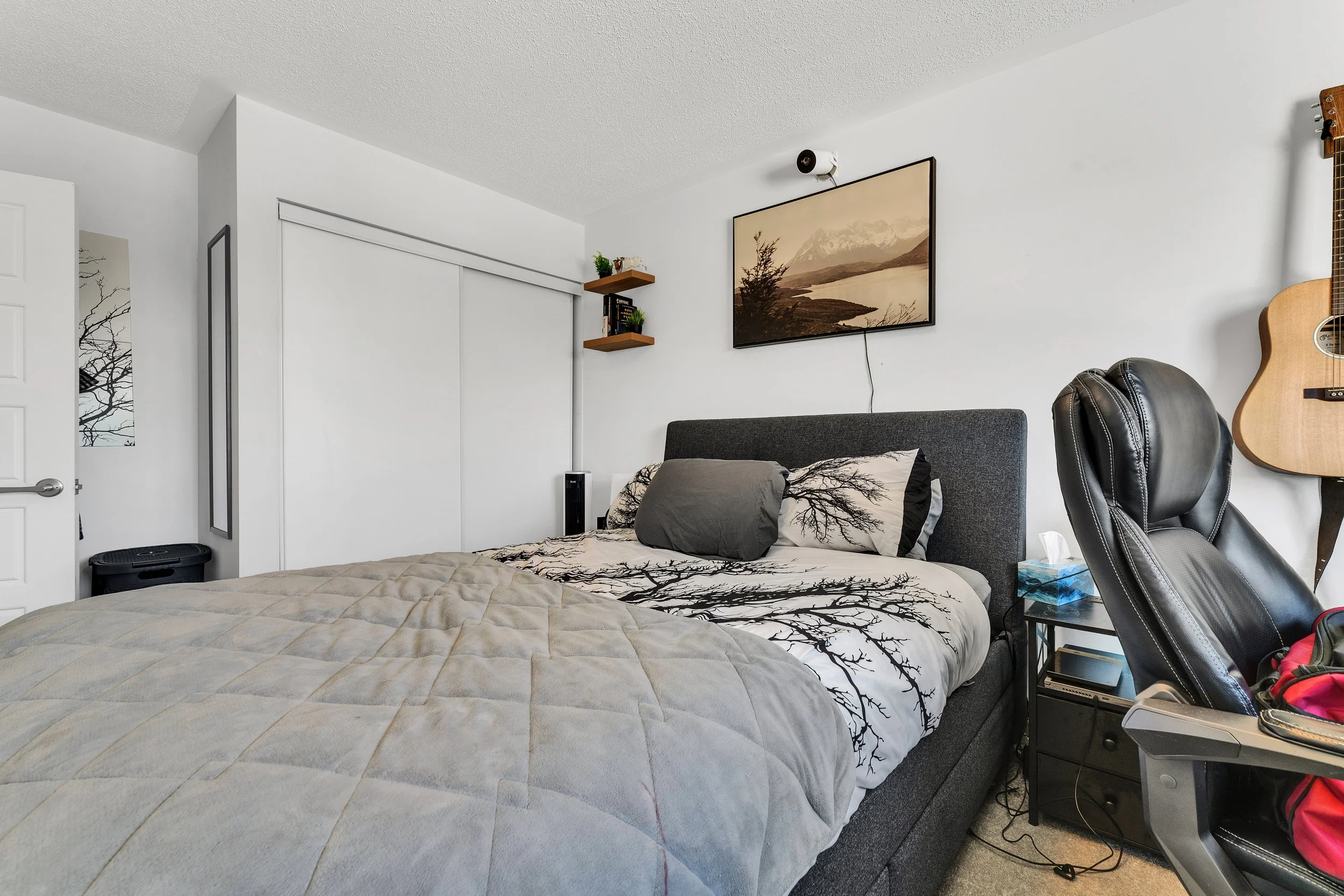 A bedroom with a bed, black headboard, patterned bedding with tree branches, a gray pillow, a side table with tissue box, a black office chair, a guitar on the wall, and a wall-mounted television