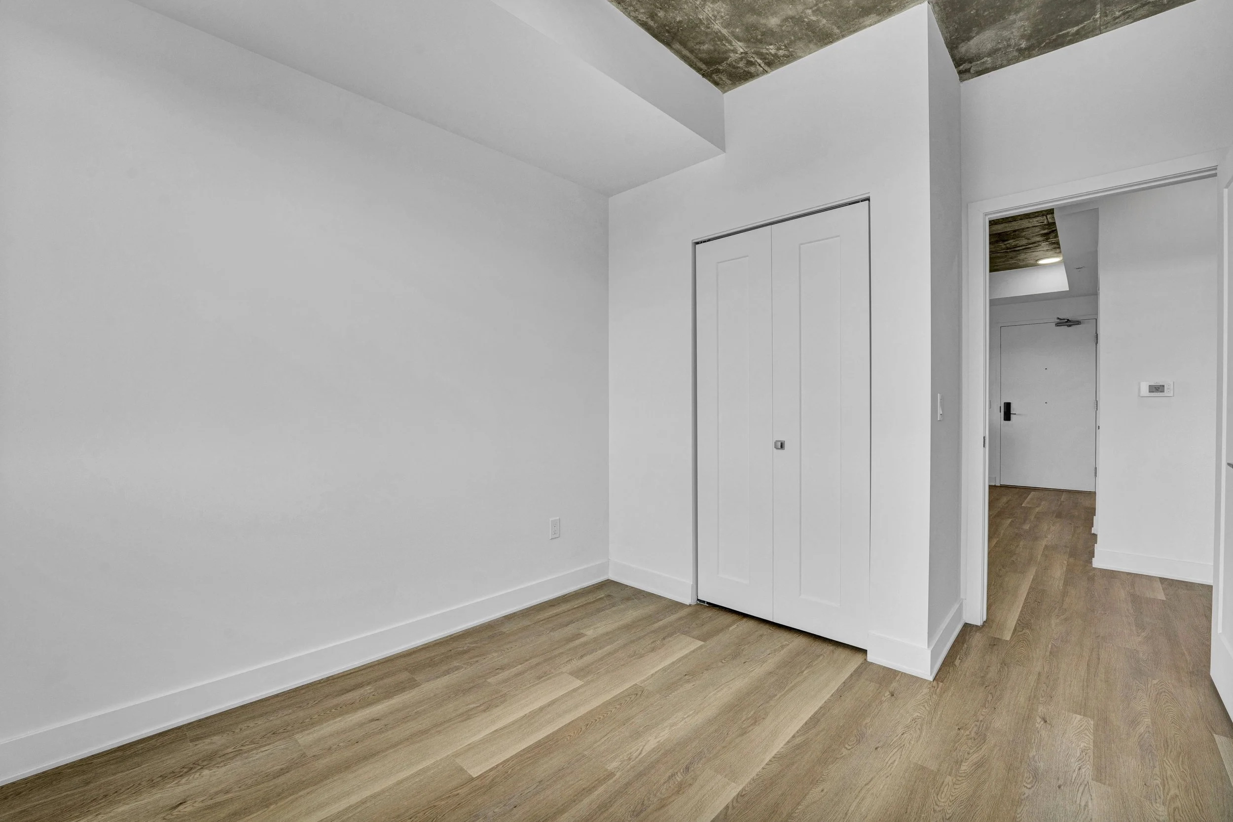 An empty room with white walls, wooden flooring, a closet with white doors, and a view into a hallway with a partial view of a ceiling with exposed concrete.