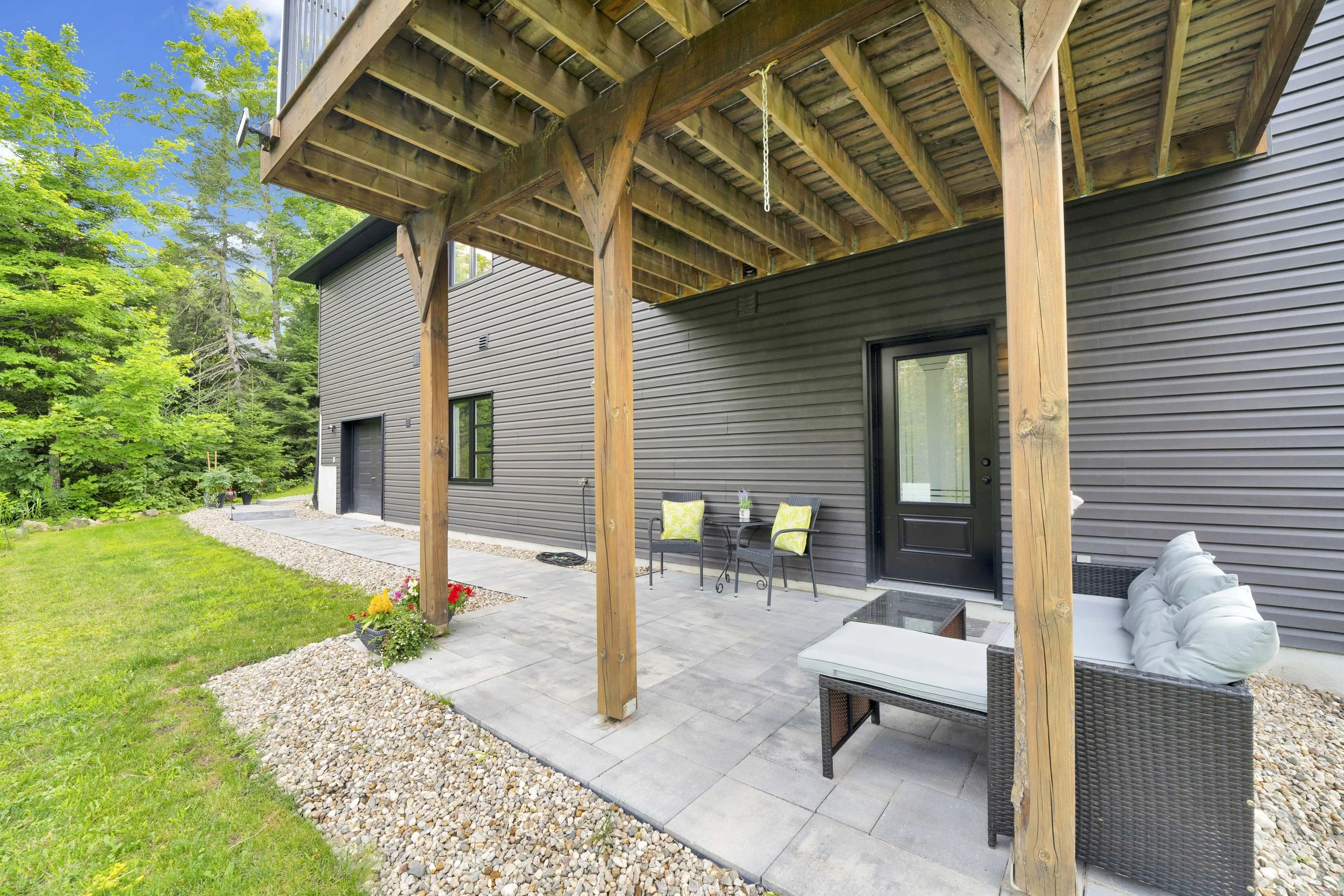 Back patio area with a black door and gray siding on the house; outdoor furniture including two chairs with yellow cushions, a coffee table, and a white outdoor sofa with pillows; wooden deck above supported by vertical wooden posts; gravel border ar