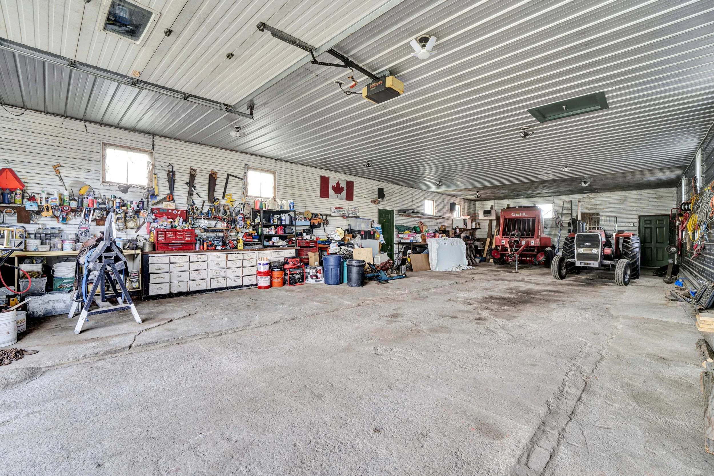 A cluttered garage with tools on shelves, a Canadian flag on the wall, and two red tractors inside.