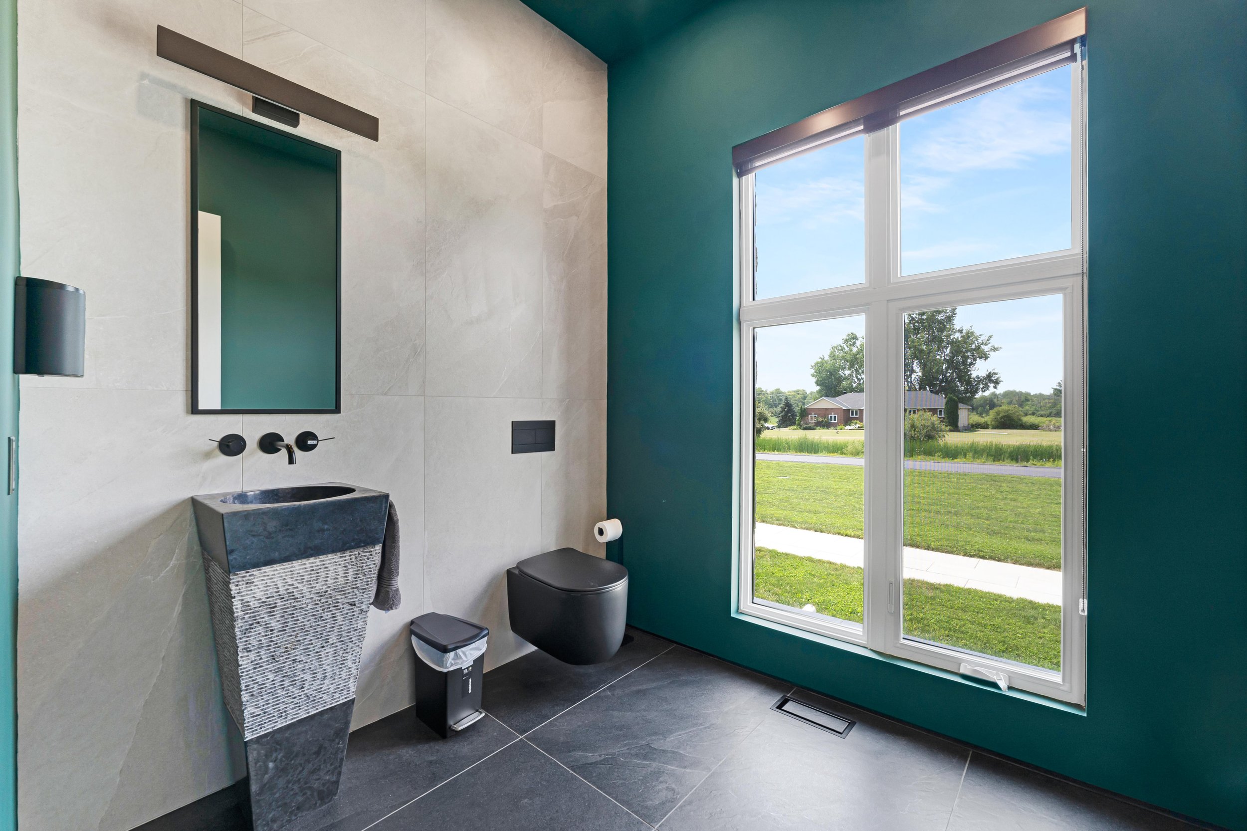 Modern bathroom with teal walls, featuring a black and gray pedestal sink, wall-mounted faucet, sleek black toilet, small trash can, and large window with outdoor view.