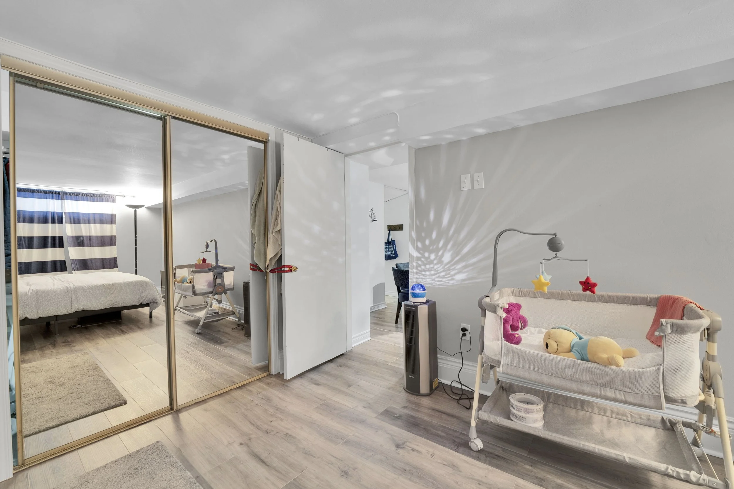 Ottawa ReThe image shows a living space with a mirrored closet, a bed, a diaper changing table with toys, a fan, and a child’s crib with stuffed animals. There is a doorway, walls, and ceiling painted white. The room has wood flooring with some rugs.