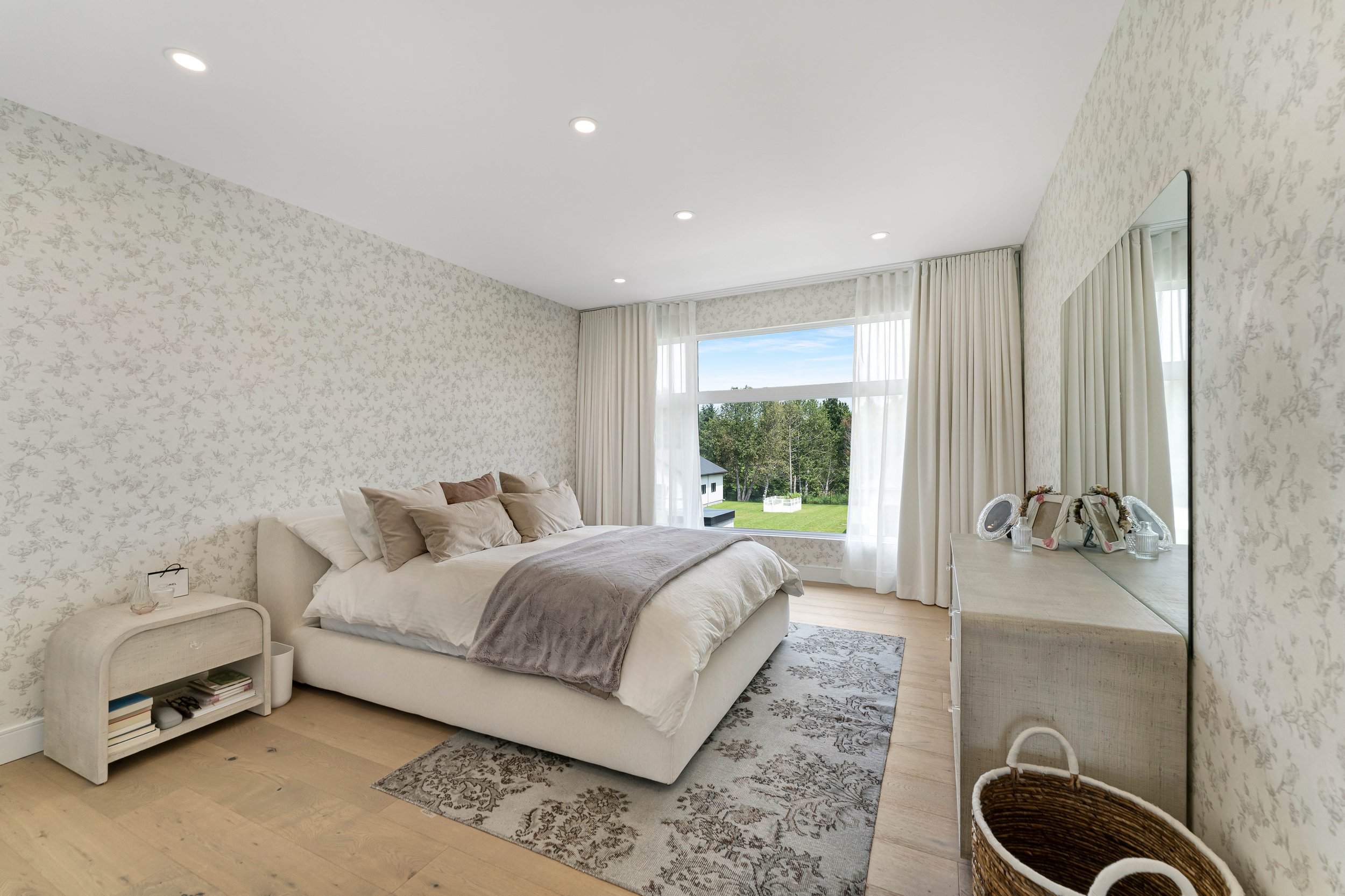 Modern bedroom interior with a large bed in the center, white and beige decor, floral wallpaper, a sizable window with view of greenery, a dresser with a mirror, and a woven basket on the floor.