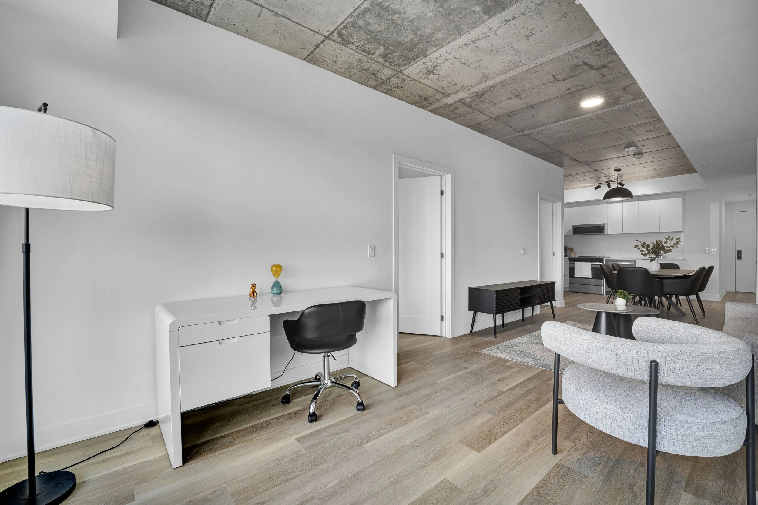 Modern living space with white walls, wooden flooring, minimalist furniture, and a concrete ceiling. Contains a white desk with a black office chair, a gray upholstered armchair, and a dining area with black chairs and a round table. Decor includes a