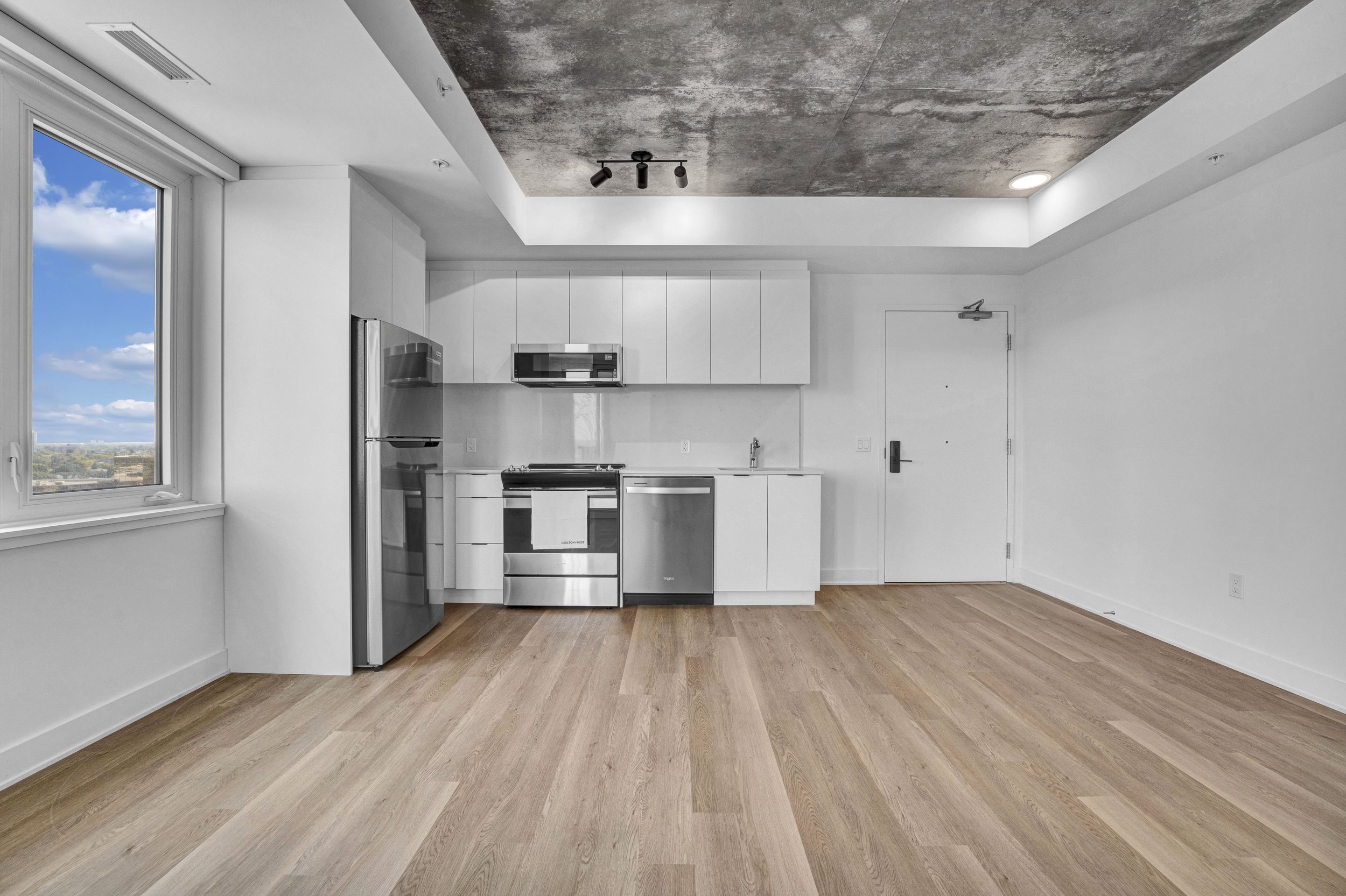 Empty modern kitchen with white cabinets, stainless steel appliances, hardwood floors, a large window with a city view, and a concrete ceiling.
