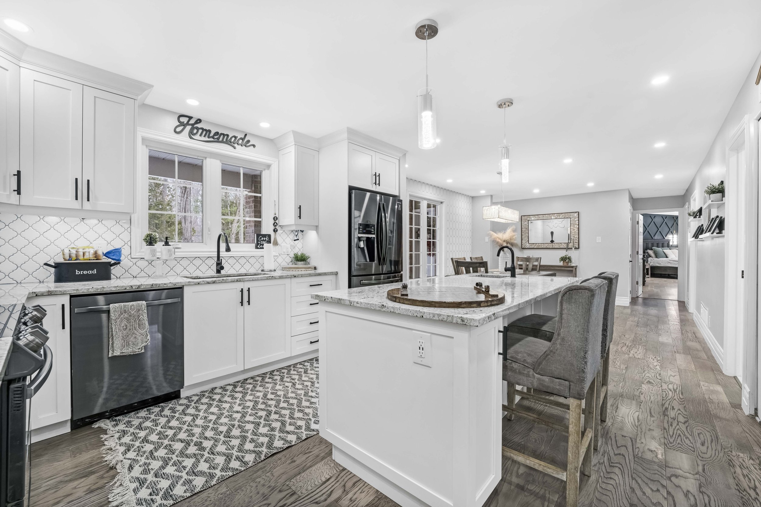 Bright, modern kitchen with white cabinets, granite countertops, and a black refrigerator, featuring a kitchen island with bar stools and a view into a dining and living area.