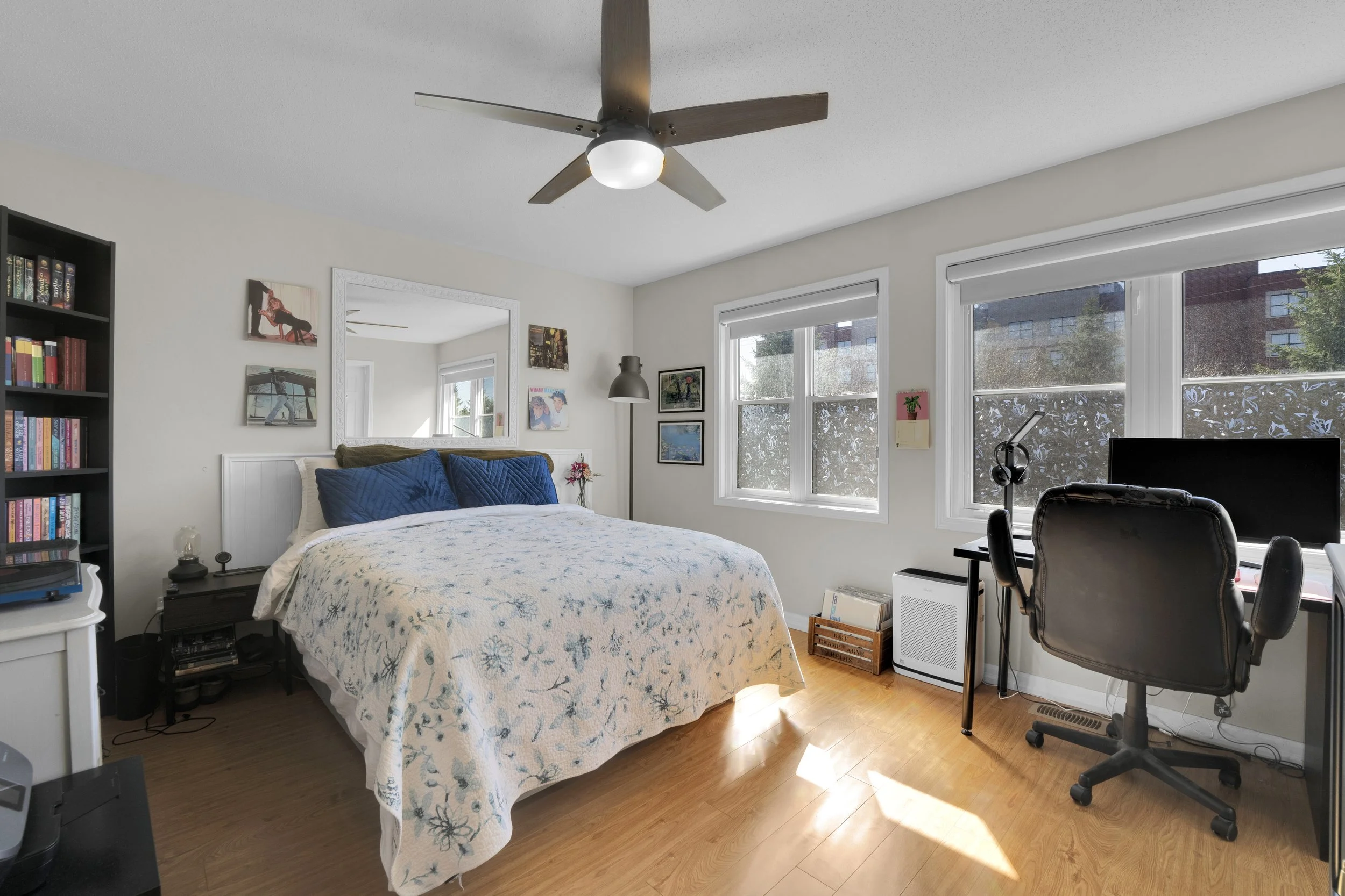 NVISION3D.ca A bright bedroom with large windows, a bed with white and blue bedding, a bookshelf filled with books, a desk with a computer and office chair, and wall art. A ceiling fan with wooden blades and a light fixture is mounted on the ceiling.