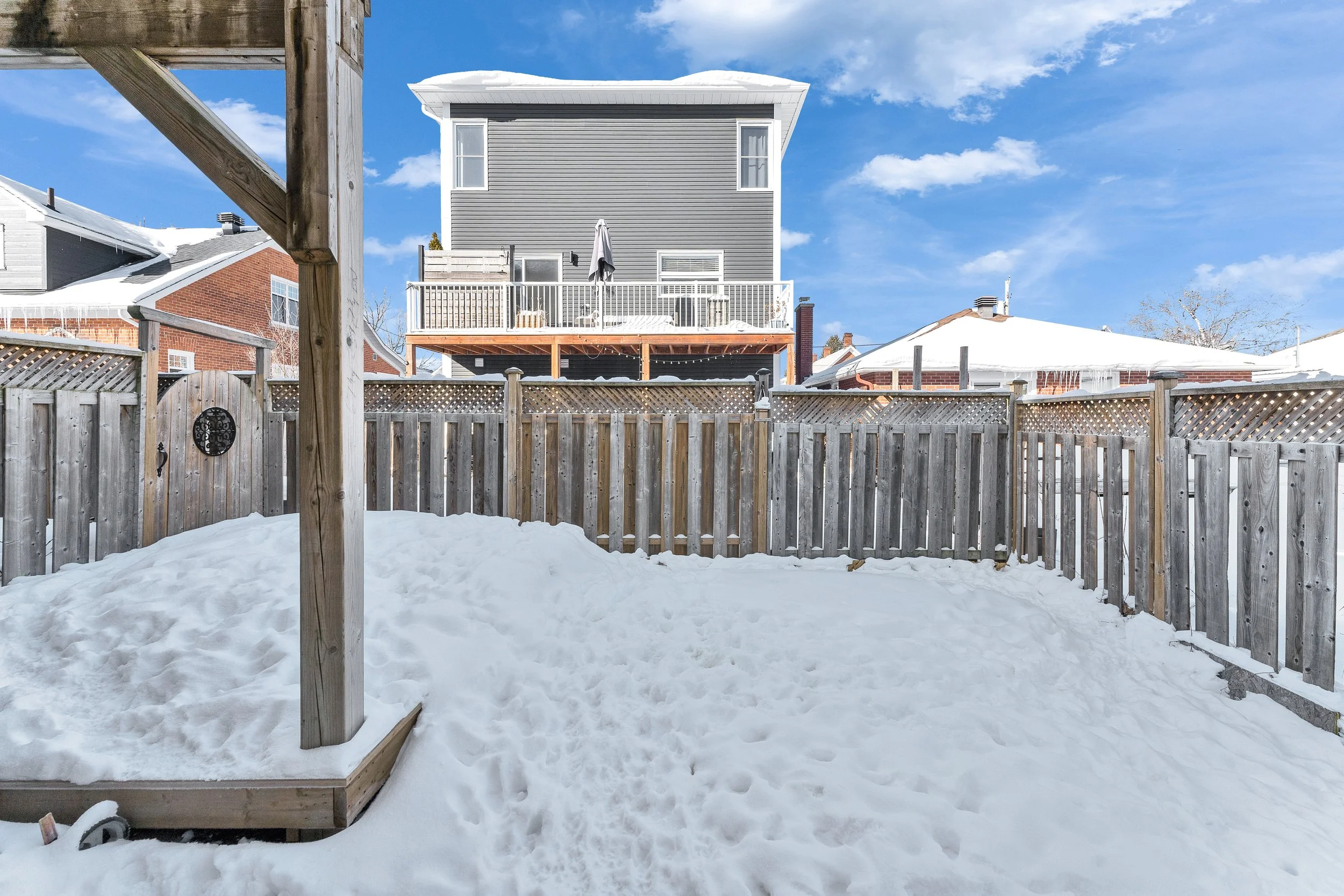Backyard enclosed with wooden fence, snow on the ground, neighboring houses with snow-covered roofs, and a blue sky with some clouds.