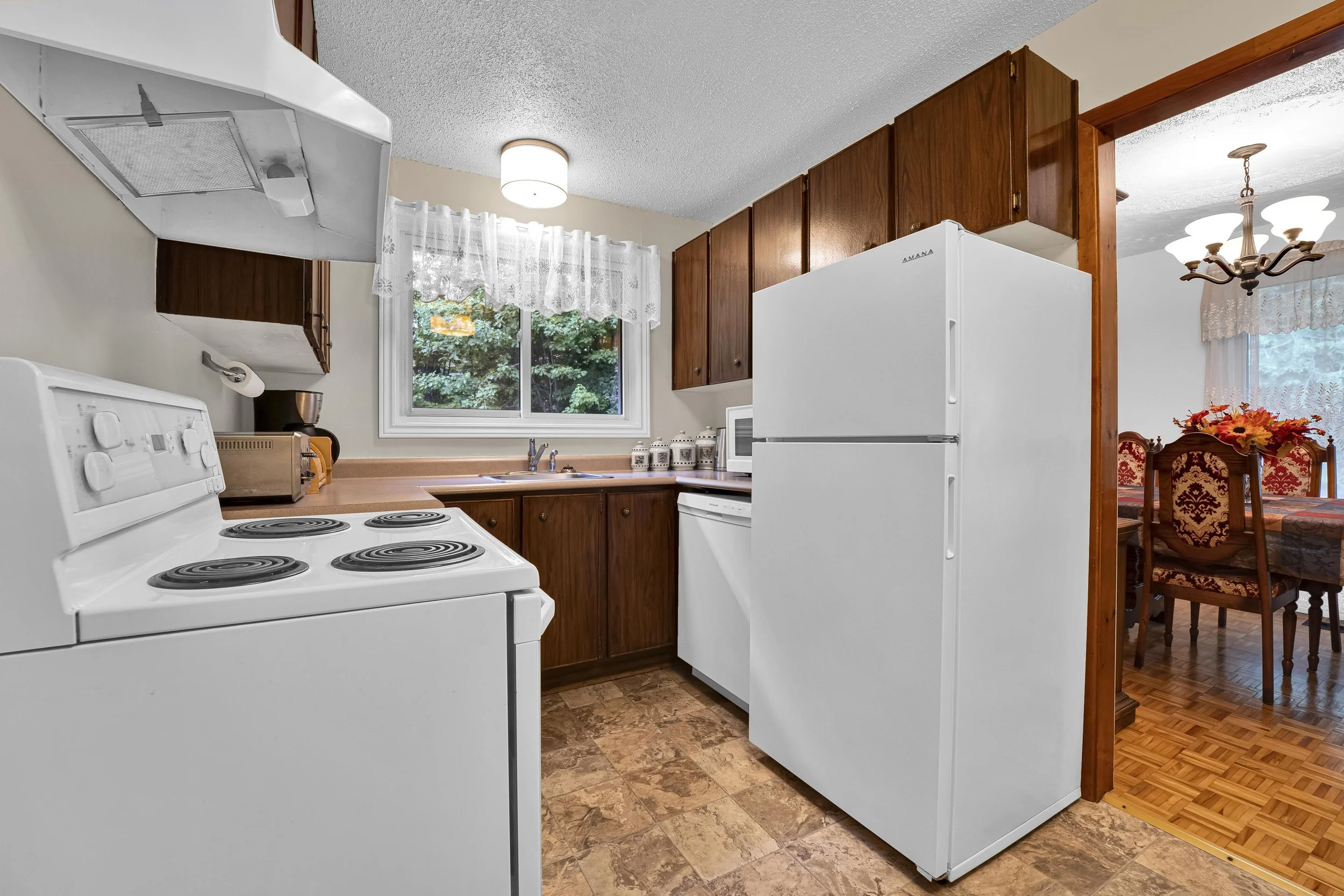 Ottawa Real Estate Photographer A kitchen with white appliances, wooden cabinets, a window with lace curtains, and a view of a dining area with a chandelier and a table with red and gold chairs.
