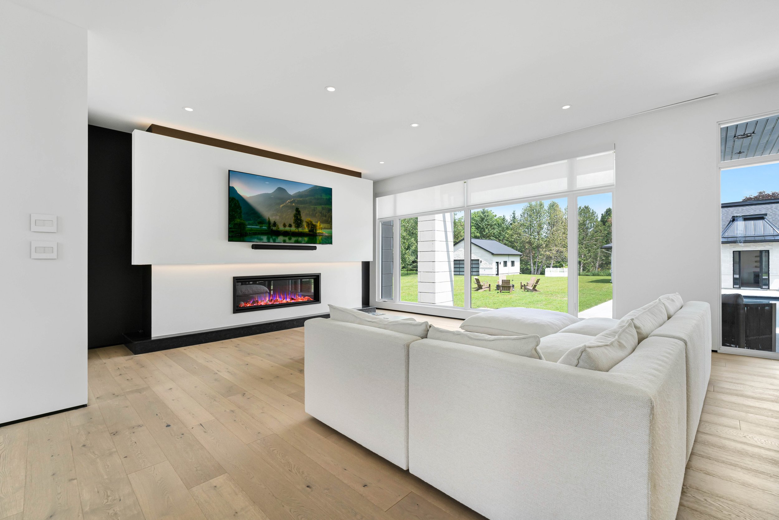Modern living room with white sectional sofa, wall-mounted TV, electric fireplace, light wood flooring, and large windows overlooking a green lawn and trees.