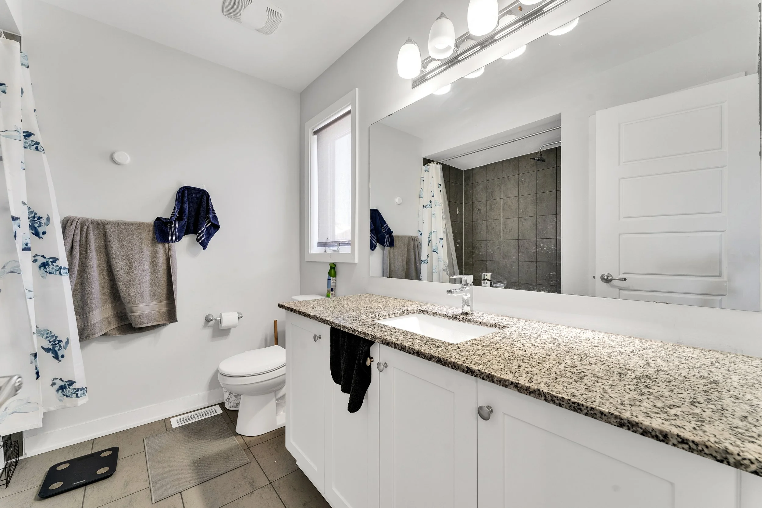 NV3D Real Estate Photography Ottawa  Bathroom with white cabinets, granite countertop, large mirror, and vanity lights. Shower area with dark tiles, toilet, window, and towel hooks with towels. Scales and bathroom mat on tiled floor.