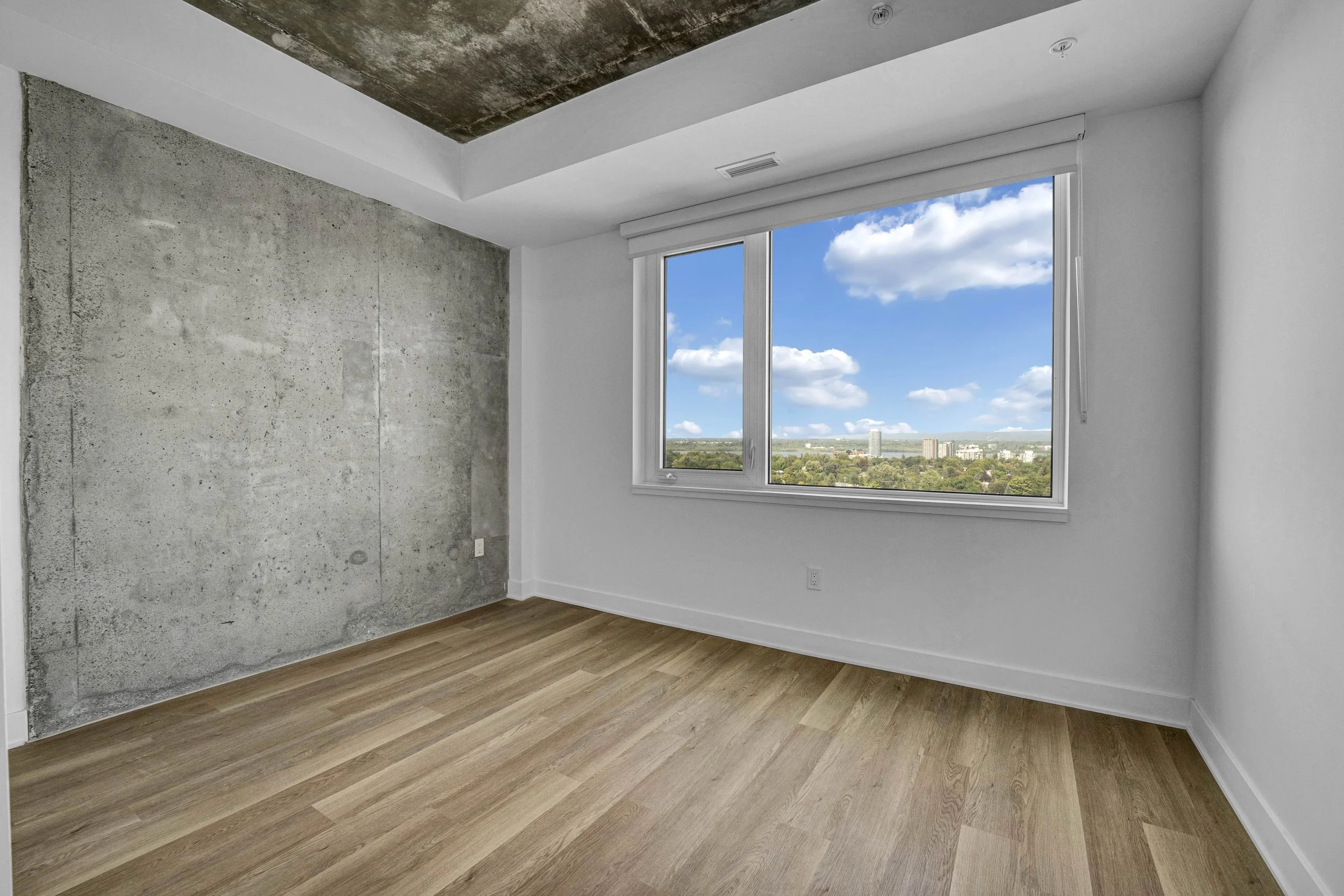 An empty room with a large window showing a cityscape and blue sky with clouds. The room has white walls, a concrete accent wall, wood flooring, and a ceiling with built-in lights.