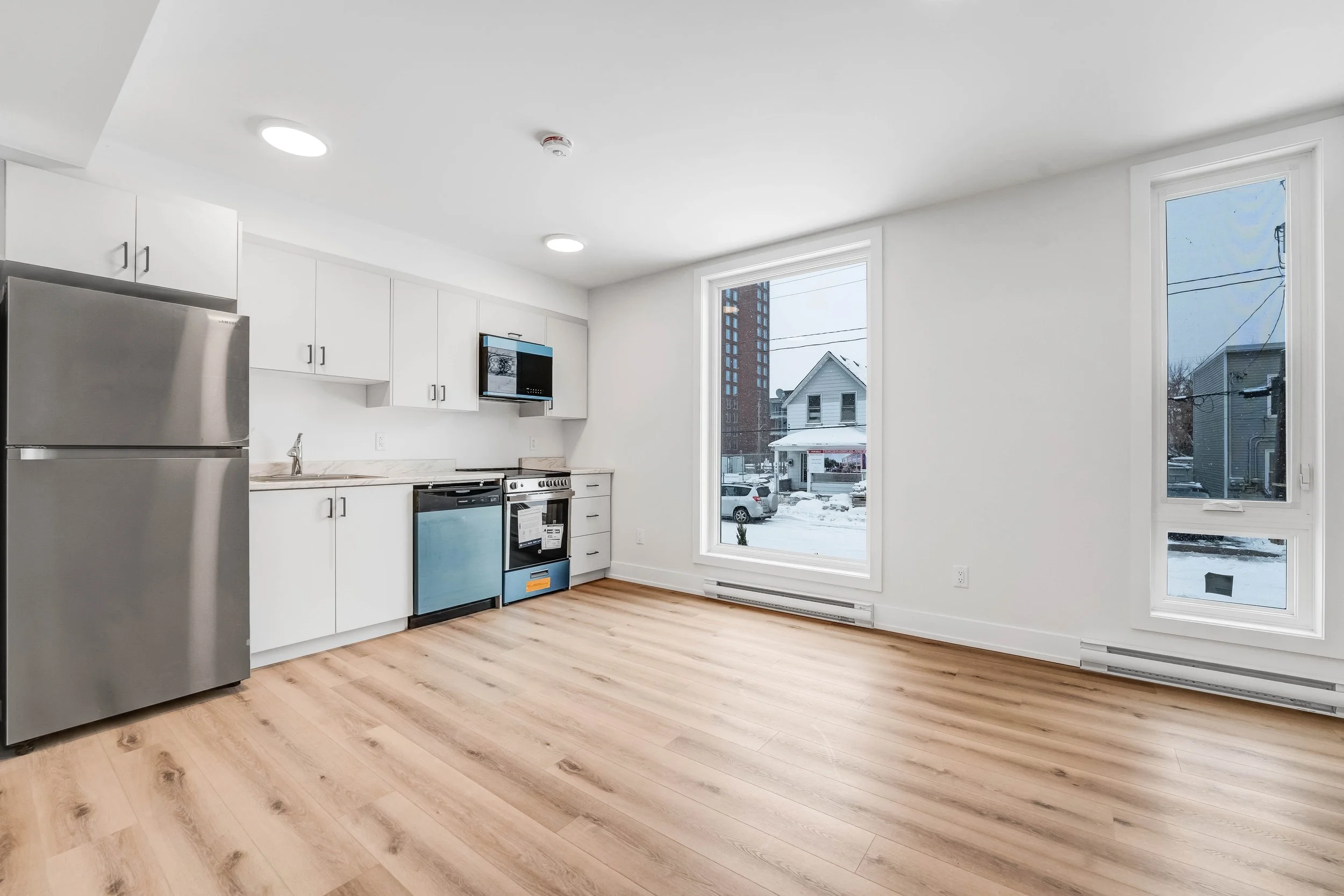 Empty modern kitchen with white cabinets, stainless steel refrigerator, stove, microwave, and hardwood floors, large window showing snowy street scene.