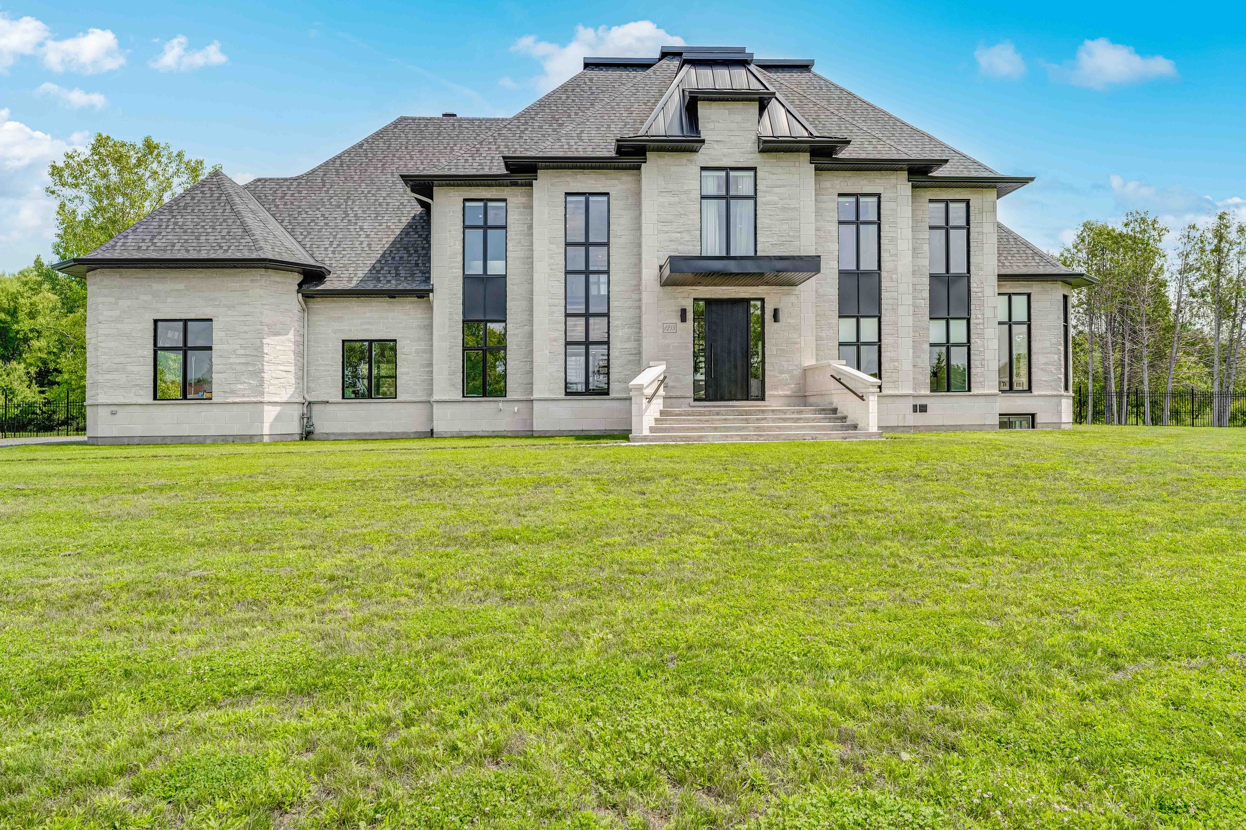 Large modern house with a gray roof, white stone facade, and multiple tall windows, situated in a grassy area with trees in the background.