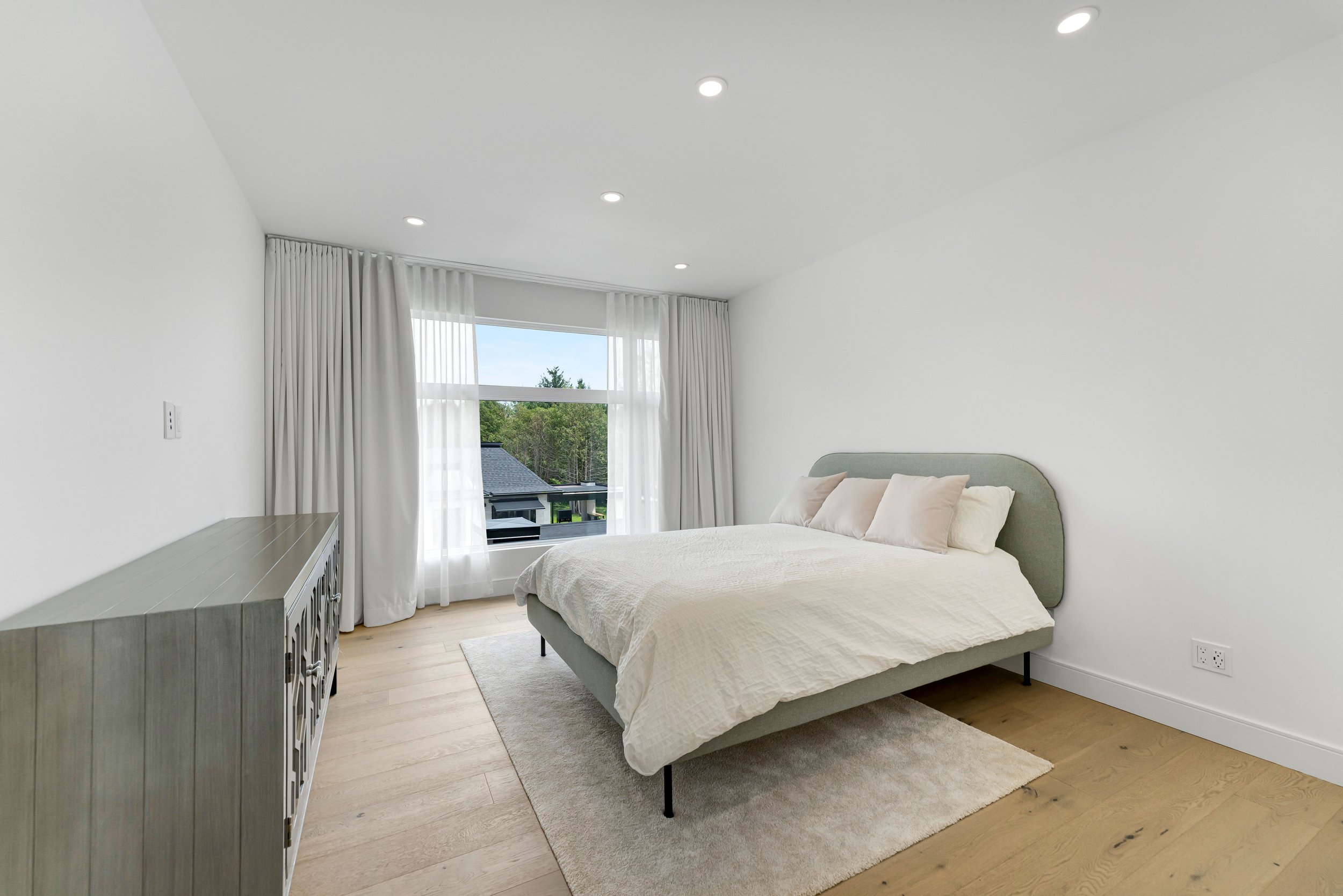 Modern bedroom with a bed, white bedding, and cushions, light wood flooring, a wooden dresser, large window with white curtains, and recessed ceiling lights.