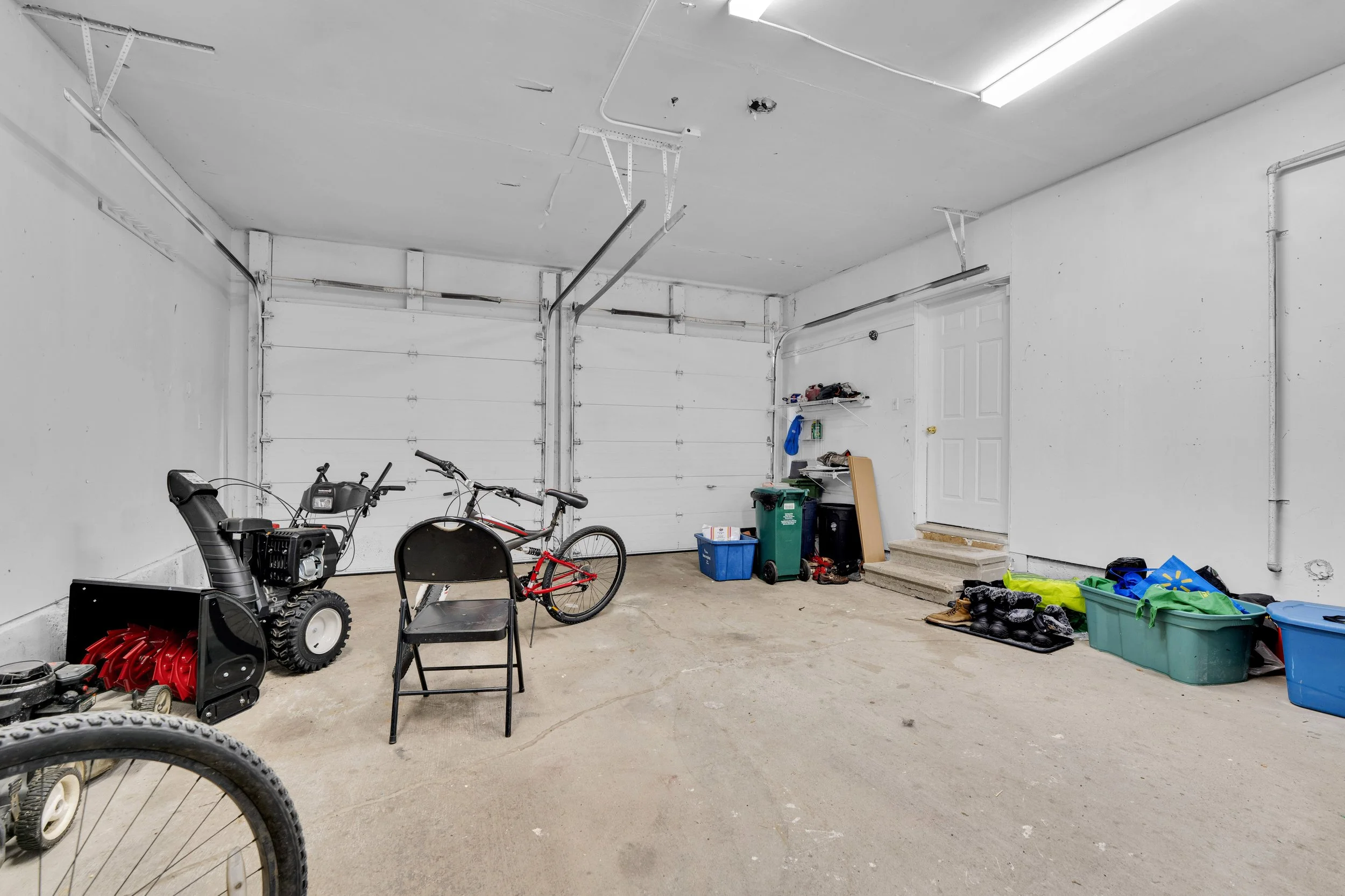 OttawA garage interior with various tools, bicycles, storage bins, and lawn equipment scattered around. The garage has a white ceiling and walls, a concrete floor, and a white roll-up garage door. There are stairs leading to a door on the right side.