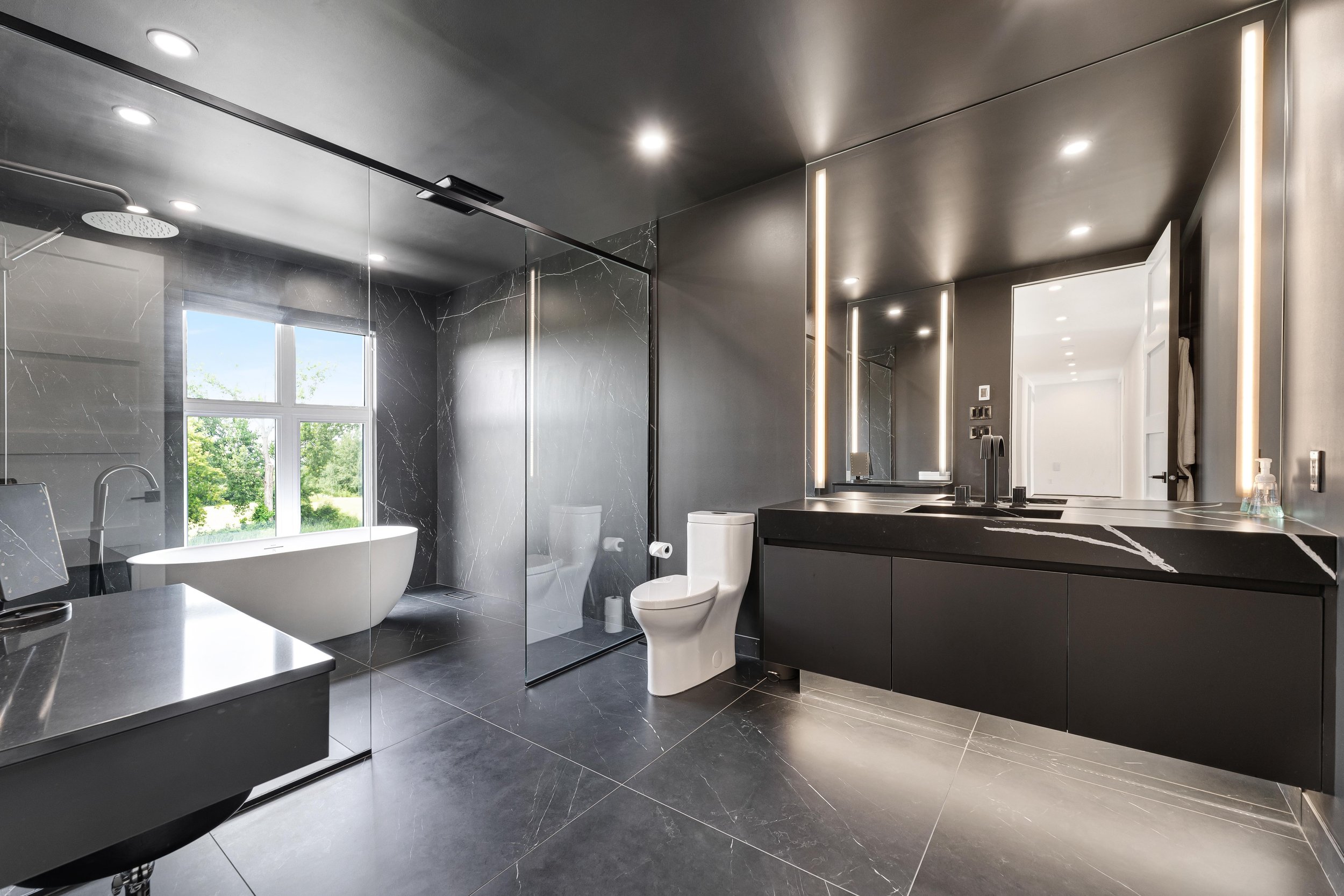 Modern bathroom with black marble finishes, featuring a freestanding bathtub, walk-in shower, toilet, large mirror, double sink vanity, and ceiling lights.