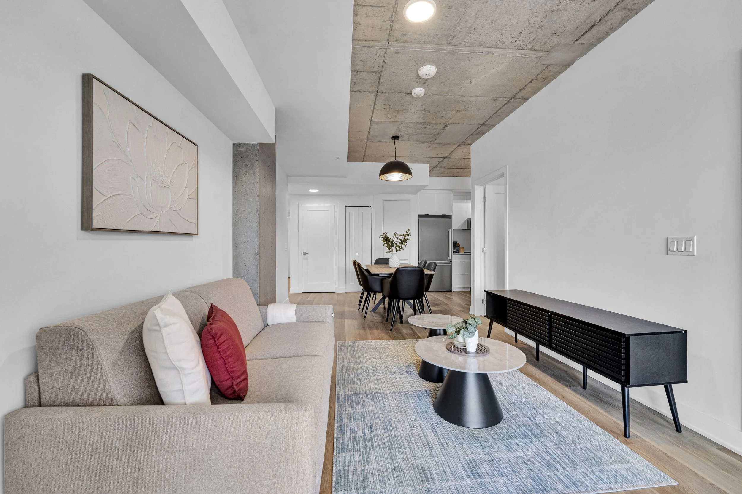 Modern living room with beige sofa, white and red pillows, two round coffee tables, a abstract flower wall art, and an dining area in the background with black chairs and a wooden table, wooden flooring, white walls, industrial ceiling with concrete 