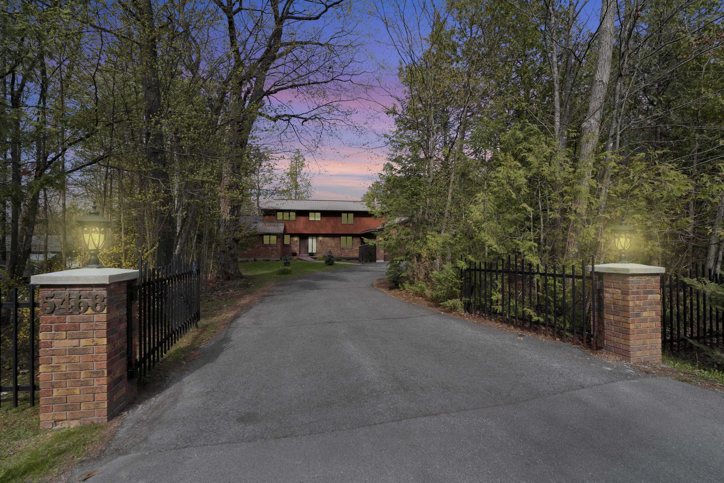 Ottawa Real Estate Media Services  View of a house entrance with a gravel driveway, surrounded by trees and a black metal fence, with a pink and purple sunset sky in the background.