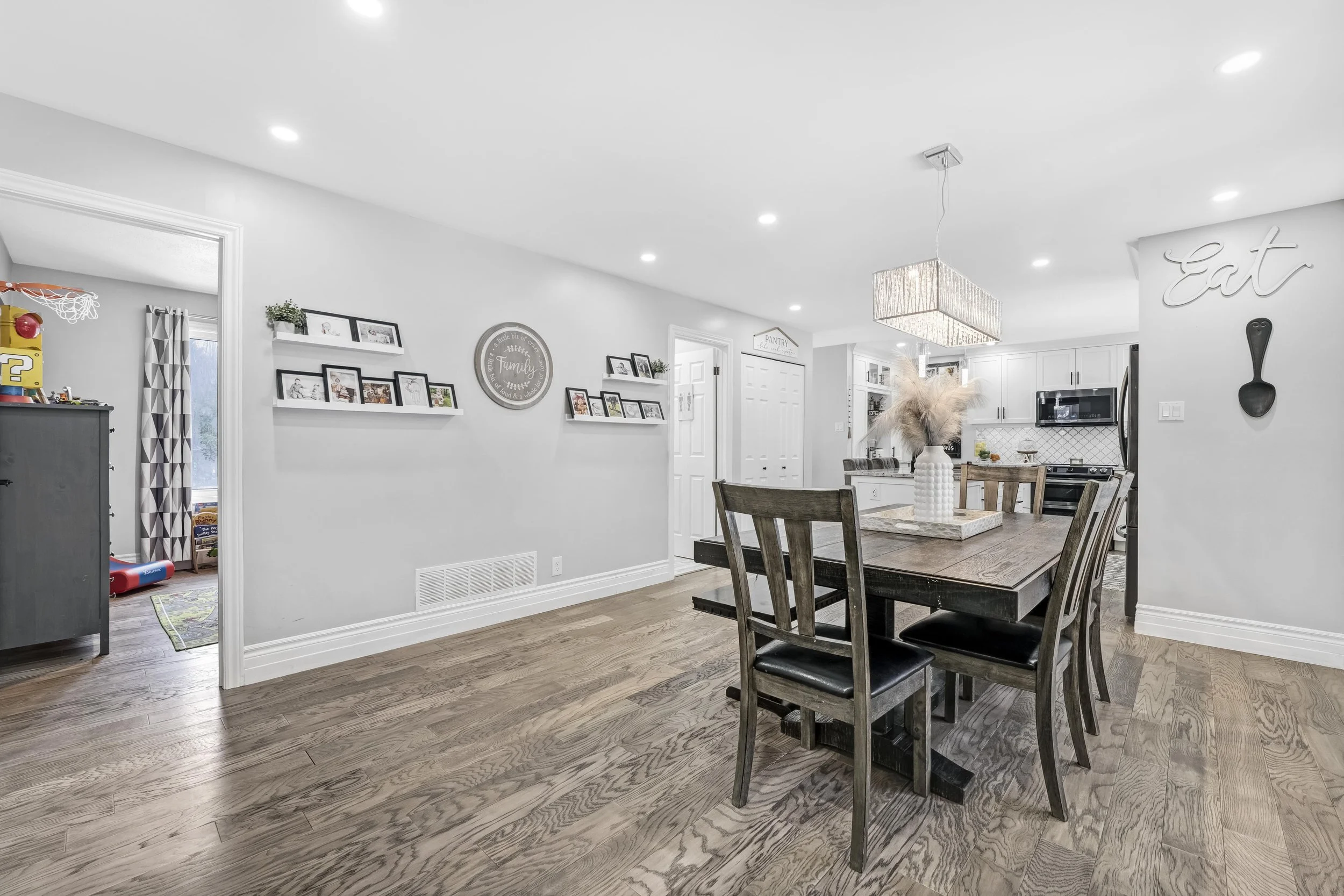 Open-concept living and dining area with white walls, wooden flooring, a dark wooden dining table with chairs, a kitchen in the background, and decorative wall shelves with framed photos.