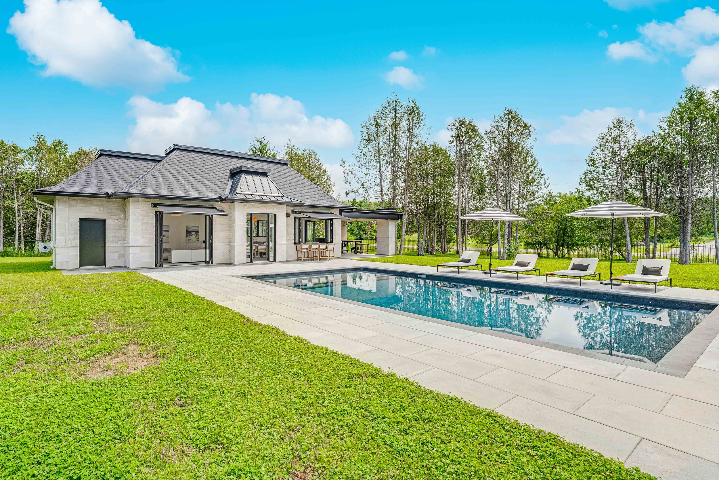 Modern pool house with a swimming pool, lounge chairs, and umbrellas, surrounded by grass and trees.