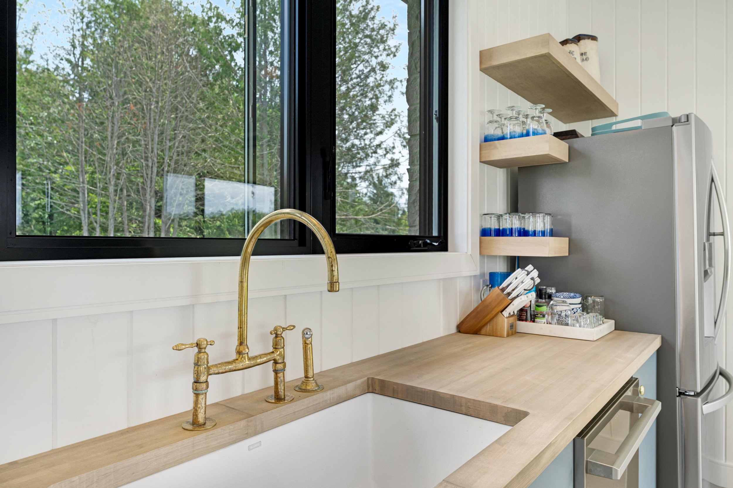 Modern kitchen with a farmhouse sink, brass faucet, wooden countertop, open shelves with glasses, cutlery holder, and a stainless steel refrigerator. Large window with outdoor view.