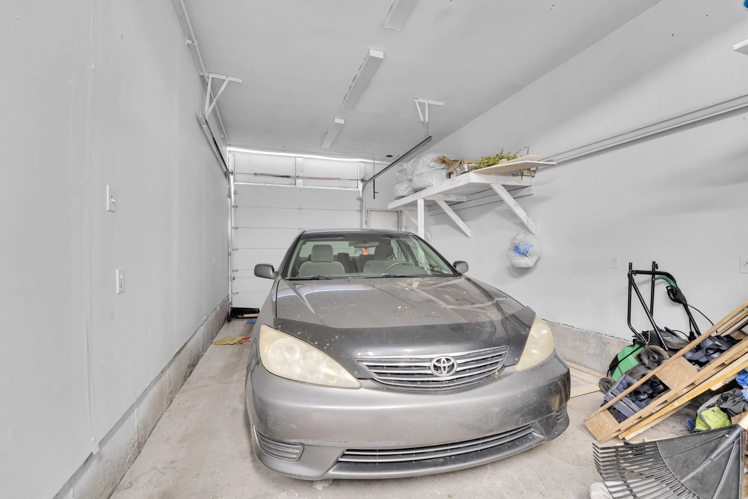 A silver Toyota sedan parked in a residential garage with a concrete floor and white walls, cluttered with miscellaneous items including a black basket, a green tool, and a wooden pallet with shoes. The garage door is closed with visible tracks and a