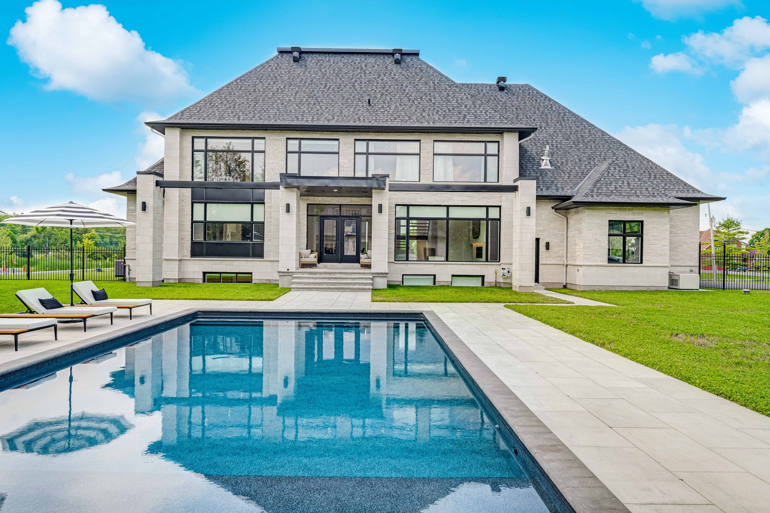 Modern luxury house with a large swimming pool and sun loungers in the backyard, surrounded by green lawn and patio area under a blue sky.
