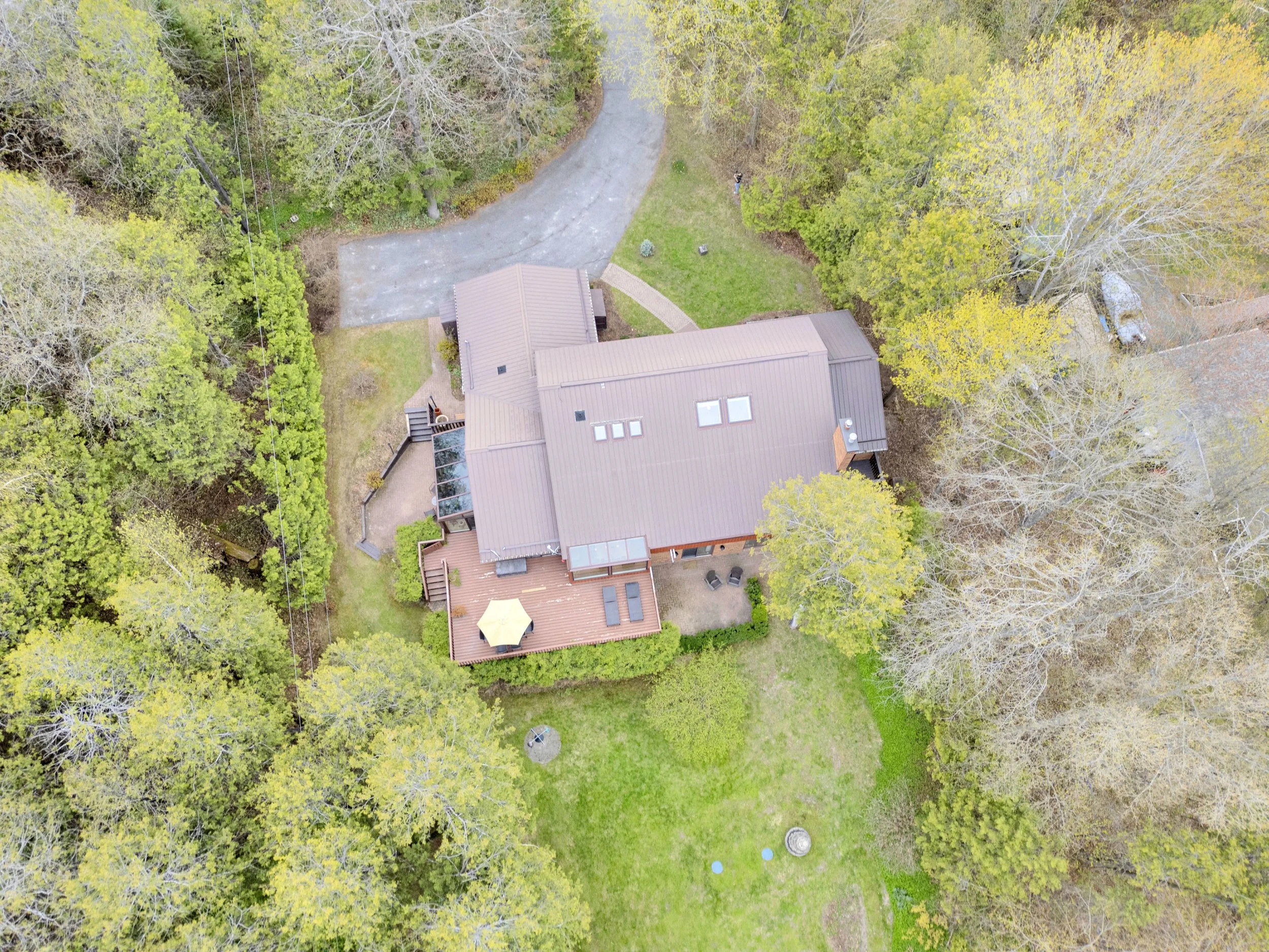 NVISION3D.ca Real Estate Media Ottawa  Aerial view of a backyard with a house, surrounding trees, a paved driveway, a deck with outdoor furniture, and a grassy lawn.