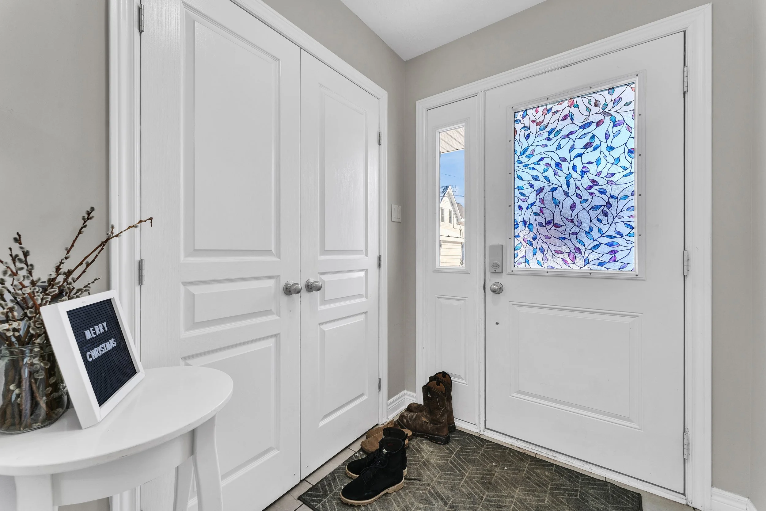 Entryway with white door featuring stained glass window, closet doors, shoes on the mat, holiday sign that says 'Merry Christmas', and a side table with a vase and dried branches.