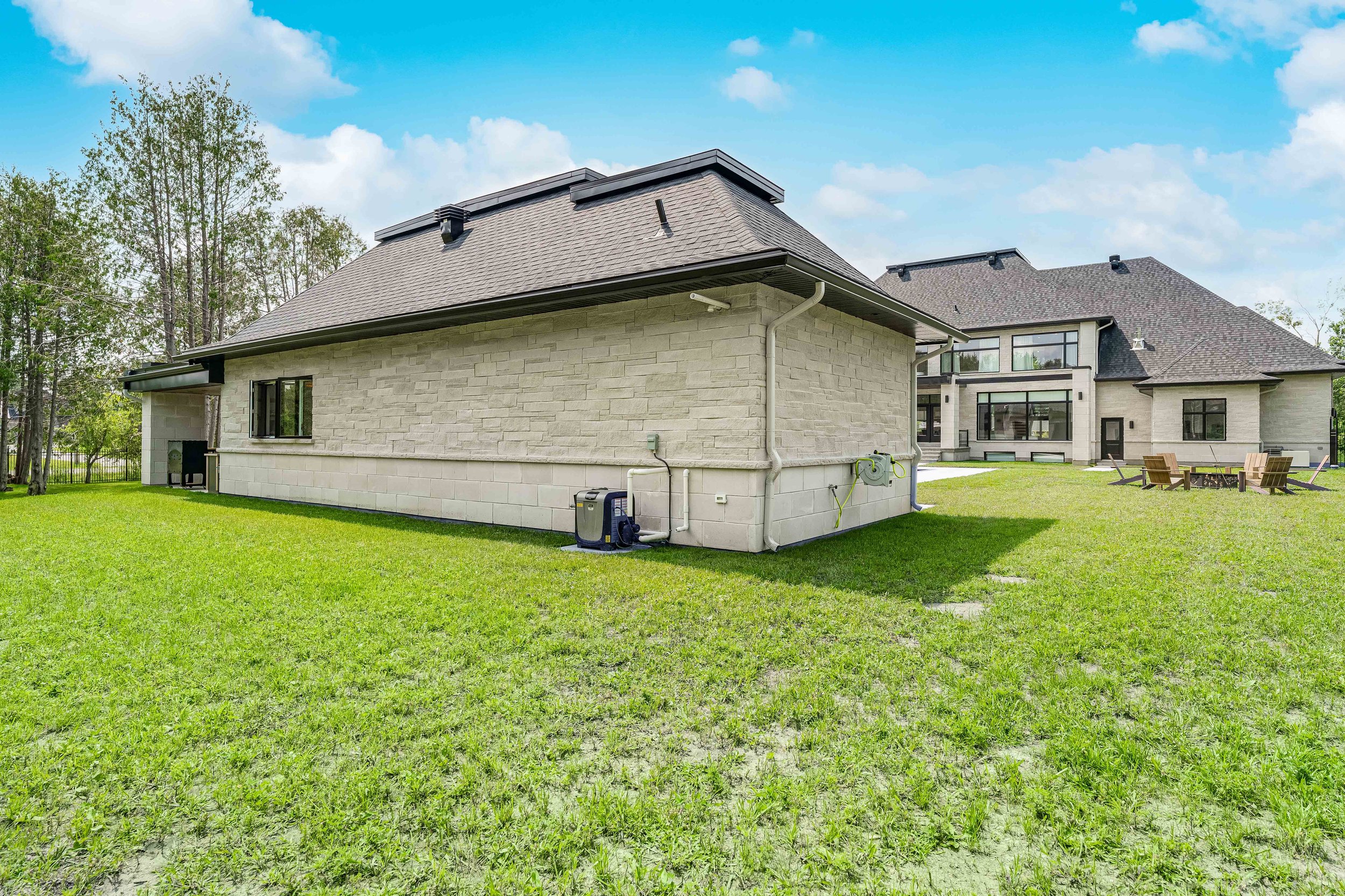 Backyard view of a large modern house with light stone exterior, manicured lawn, wooden lounge chairs, and tall trees. The sky is clear and blue.