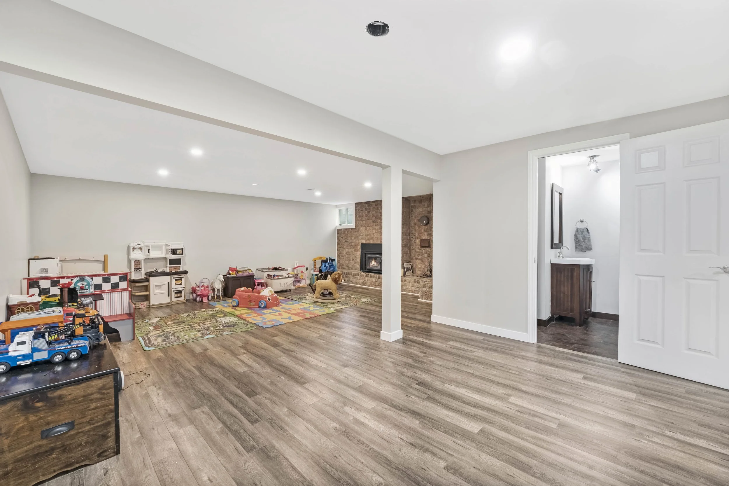 A spacious basement playroom with wood flooring, white walls, and a brick fireplace. There are various toys and a play kitchen along the left wall. To the right, a doorway leads to a bathroom with a wooden vanity and towel hanging on a ring. The ceil