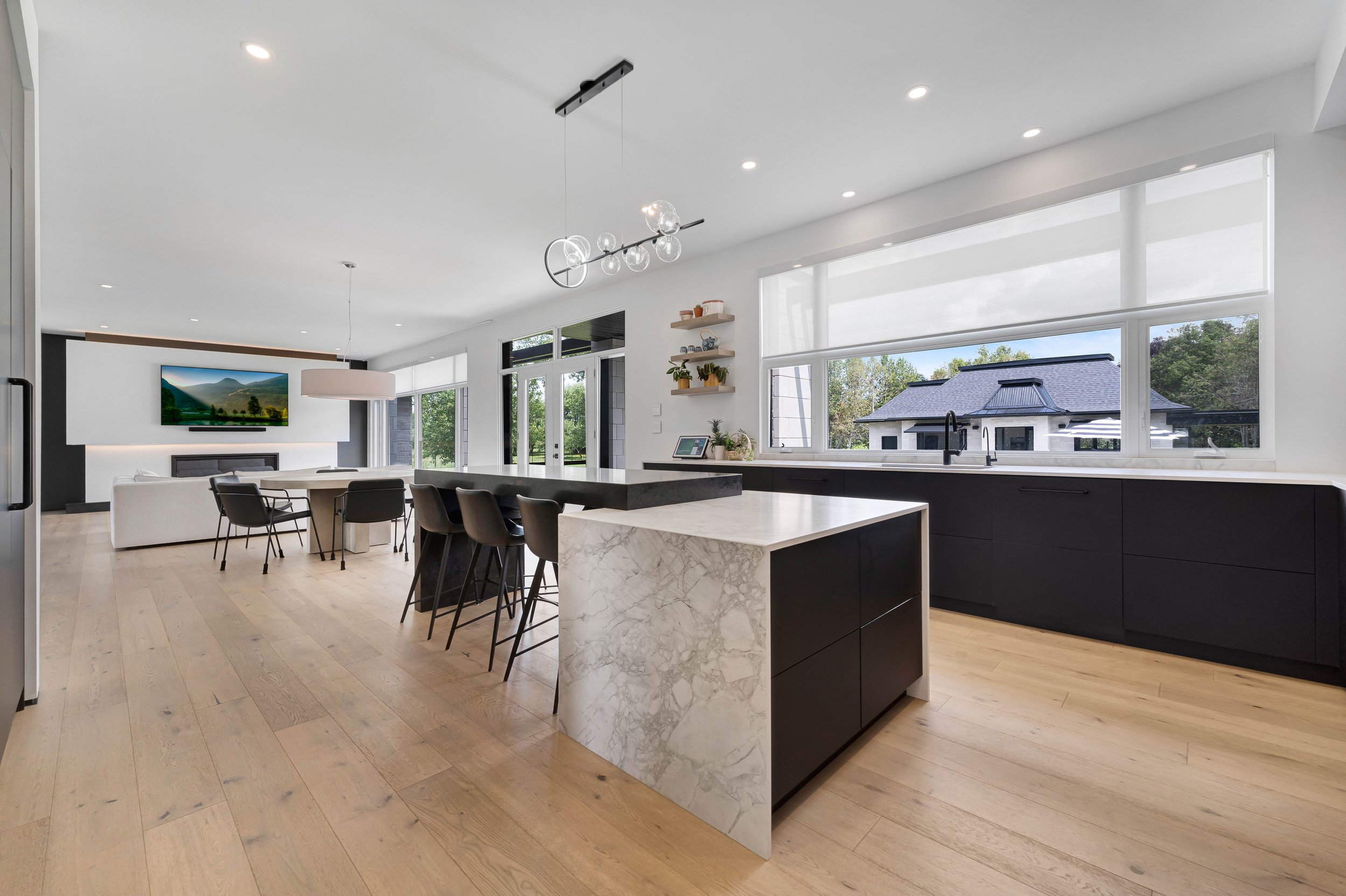 Modern open kitchen and living area with hardwood floors, large windows, dark kitchen cabinets, marble island, dining table, chairs, pendant lighting, TV, and view of outdoor space.