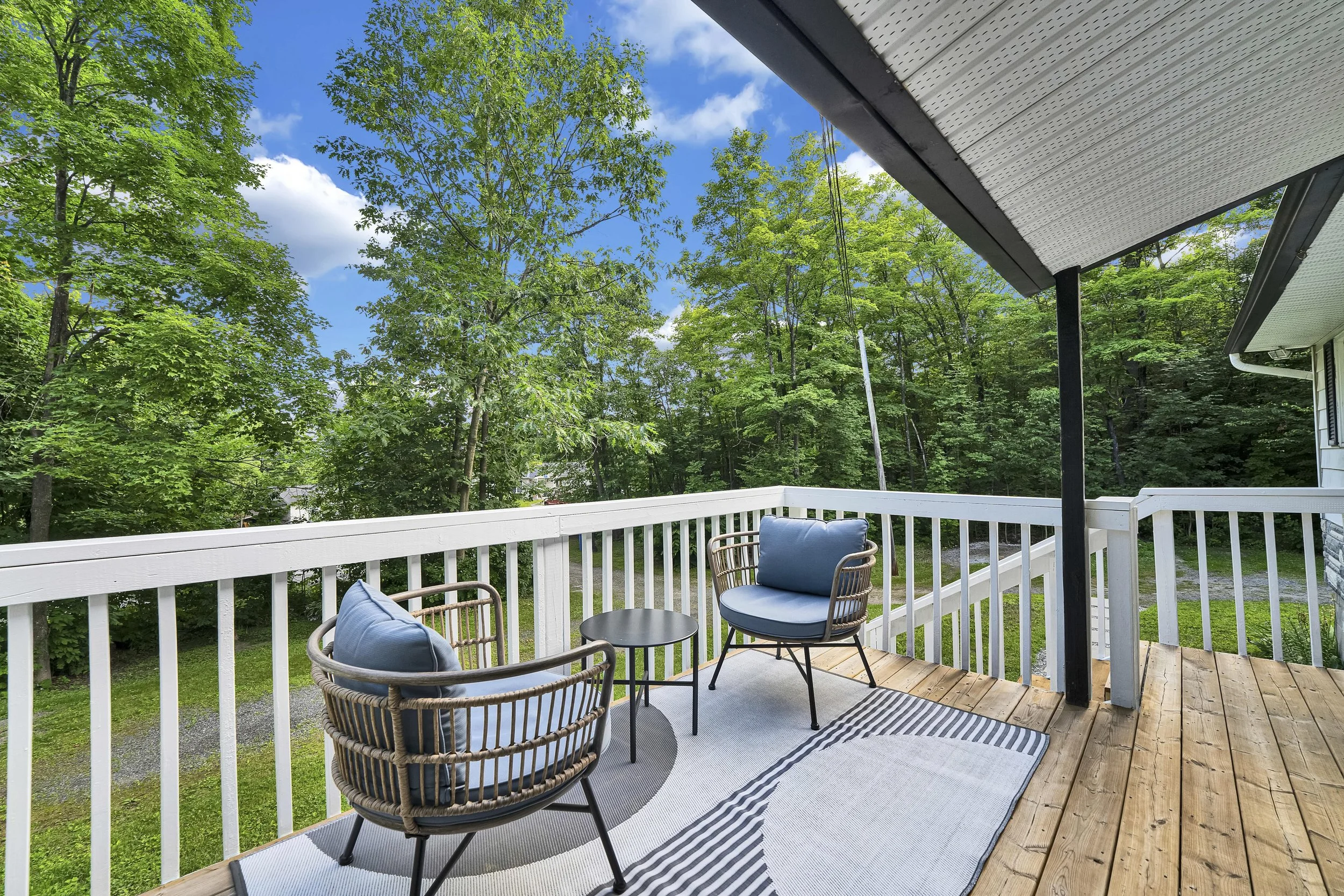 A balcony with outdoor furniture including a woven chair with cushion and a small round table on a striped rug, overlooking green trees and a blue sky with clouds. Part of the house's white railing and the roof eaves are visible.