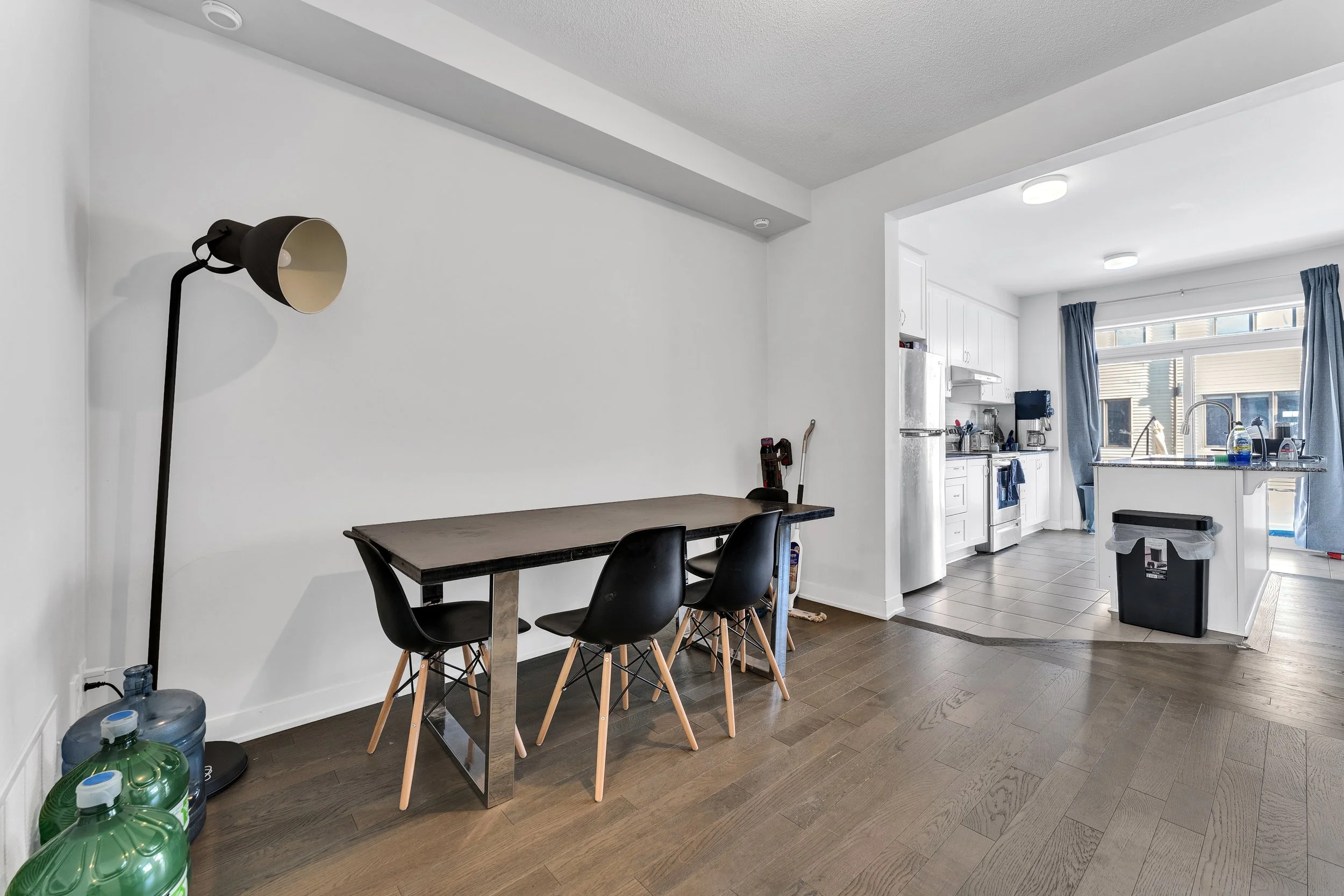 NV3D Real Estate Photography Ottawa  Empty dining area with a black dining table and four black chairs, adjacent to an open kitchen with white cabinets and a window with blue curtains.
