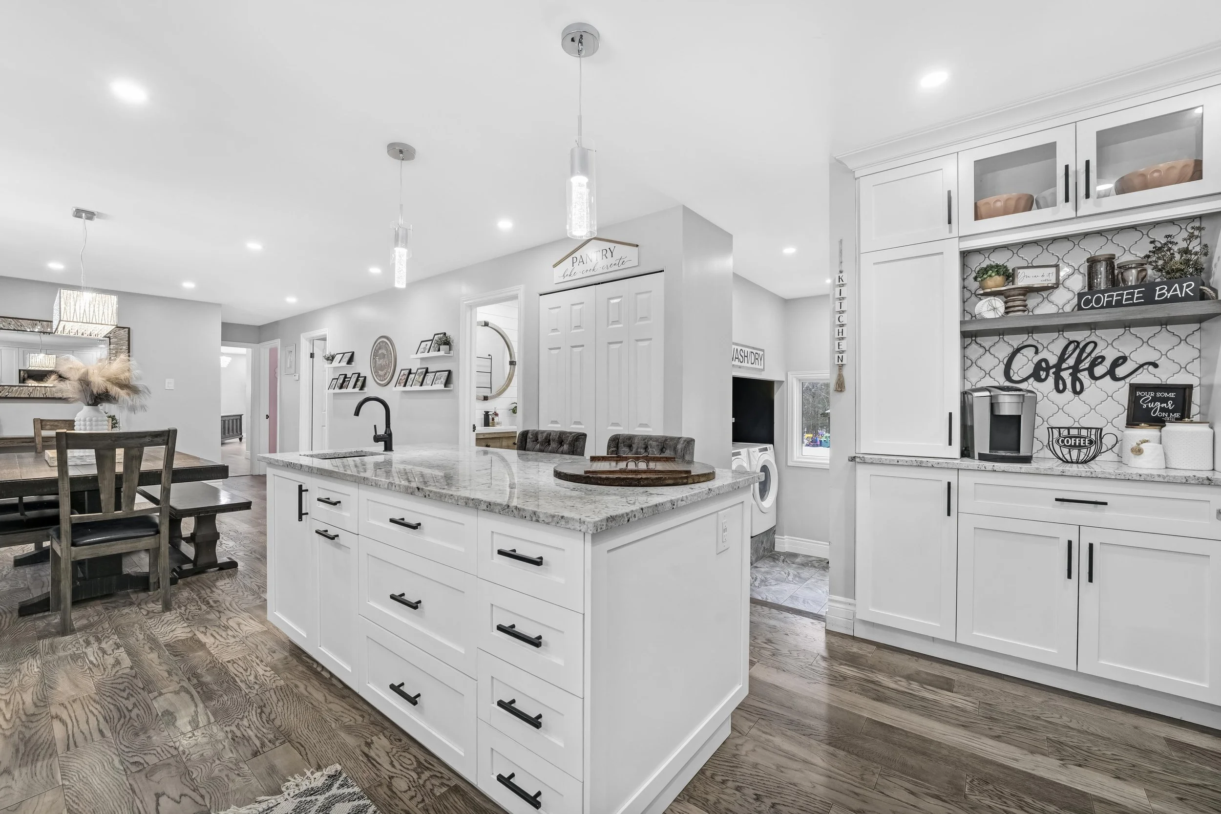 Modern open-concept kitchen with white cabinets, granite countertops, a kitchen island, and a coffee station with decor. Nearby dining area with a wooden table and chairs. Hardwood floors and recessed lighting.