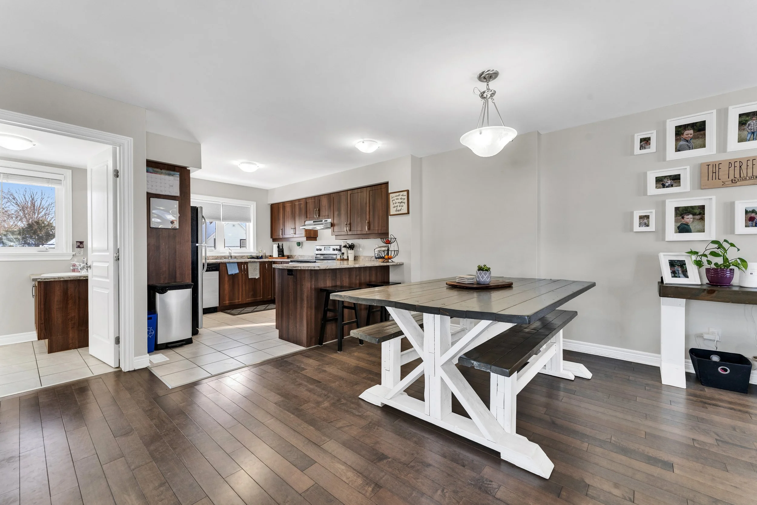 A kitchen and dining area with hardwood floors, featuring a wooden dining table with white legs and benches, brown cabinetry, white walls, and framed photos on the wall.