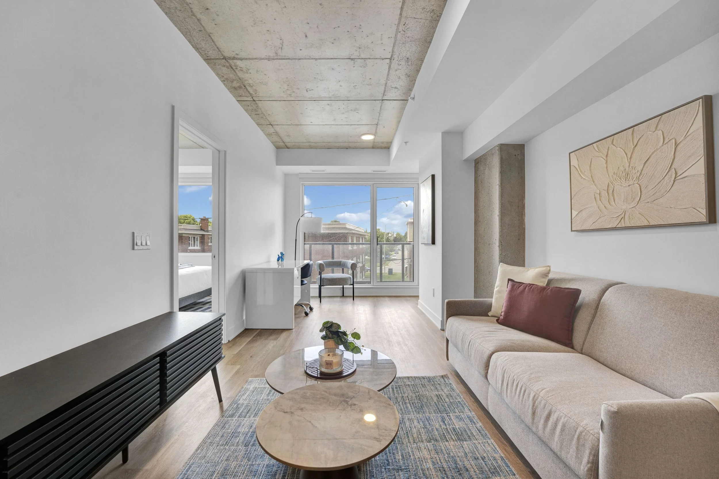 Ottawa RealA modern living room with white walls, a beige sofa with rust-colored pillows, a small round coffee table with a candle and plant, a black media console, large windows showing a neighborhood view, and a piece of floral artwork on the wall.