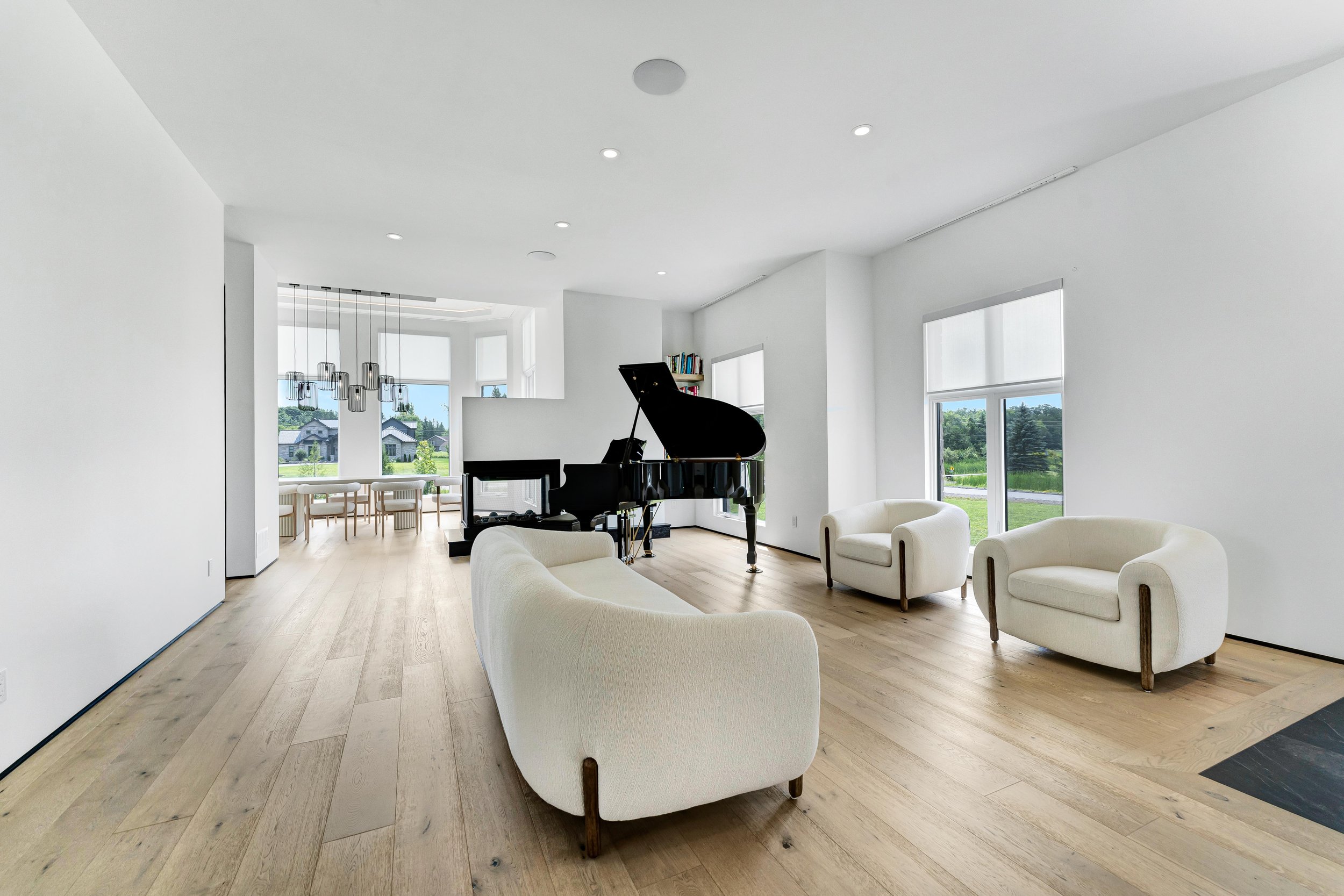 Modern living room with a grand piano, white sofas, armchairs, large windows, and a dining area with a view of greenery.
