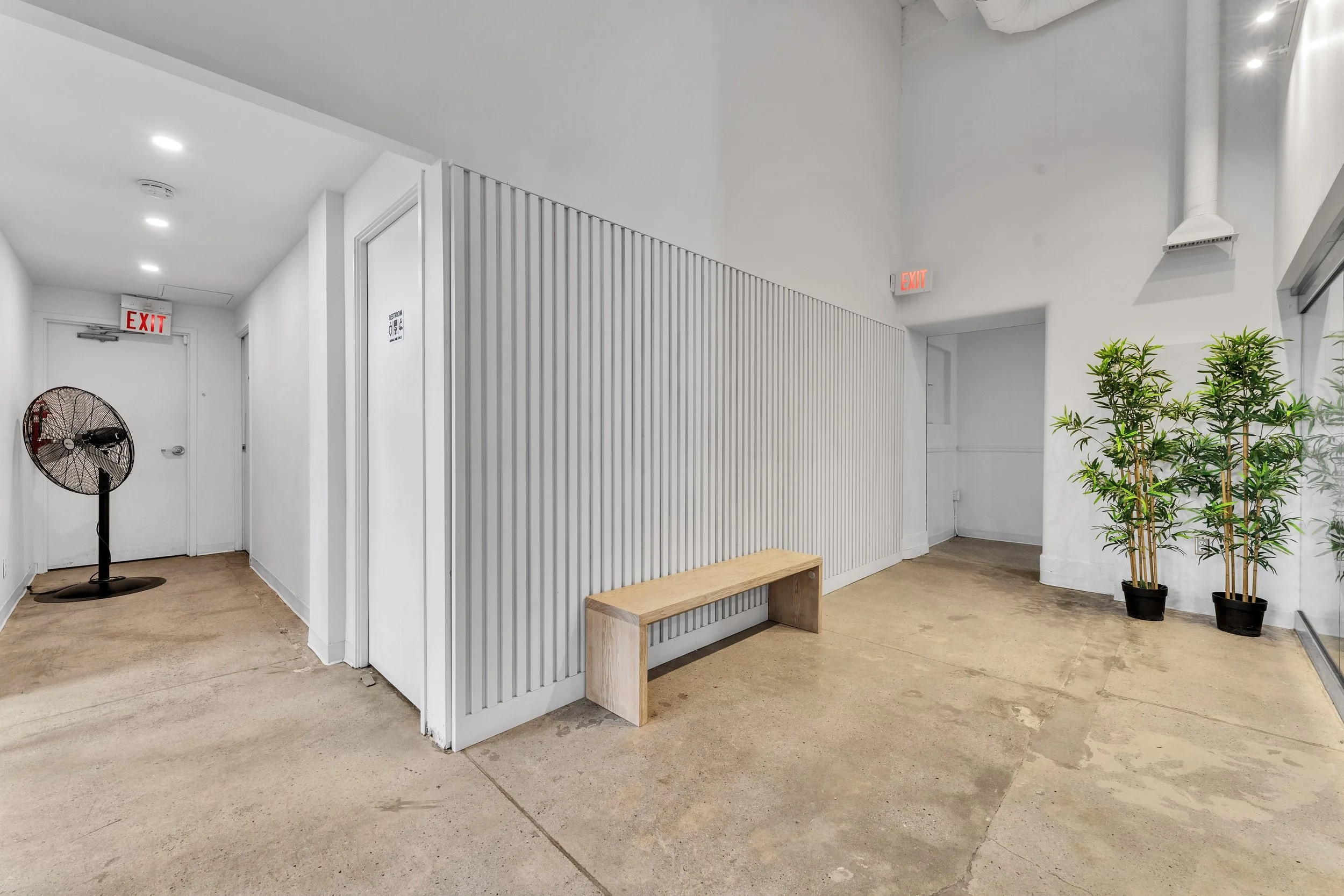 A minimalistic lobby or waiting area with a wooden bench, a potted plant, white walls, and a standing fan. There are two exit signs and two doors.