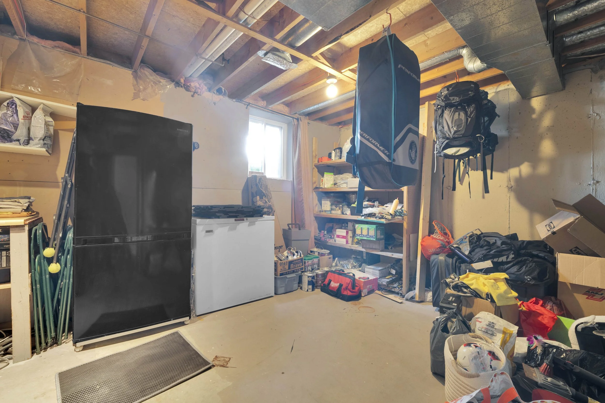 A cluttered garage with storage shelves, a black refrigerator, a small white freezer, and various boxes, bags, and outdoor gear scattered around. There is a window allowing natural light to come in and exposed insulation and wooden beams on the ceili