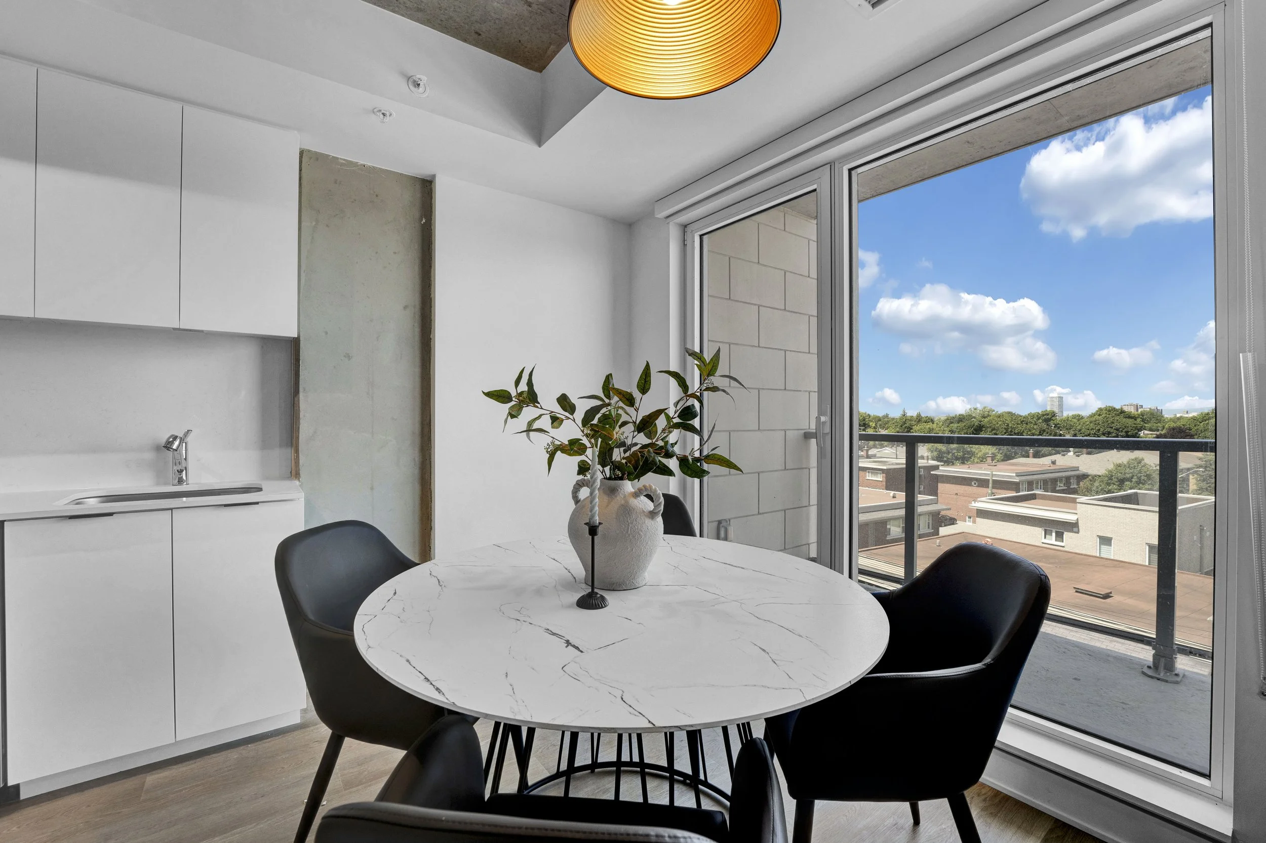 Modern dining area with a marble-top round table, black chairs, a potted plant centerpiece, large sliding glass doors with a view of a cityscape and trees, white cabinetry, and a hanging orange lampshade.