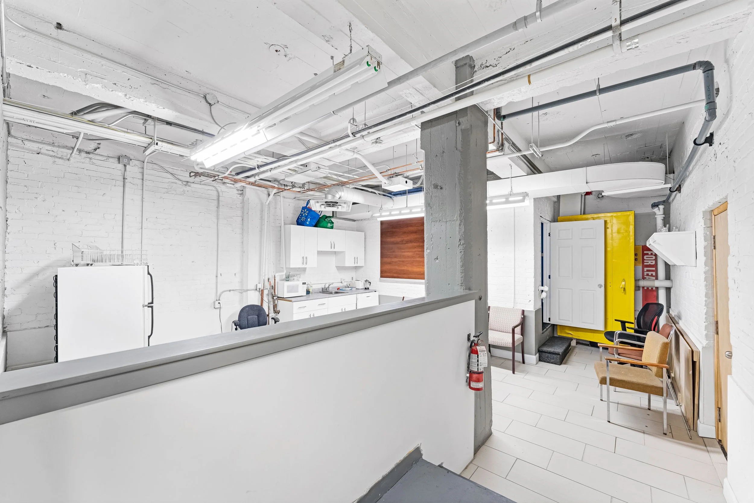 NV3D Real Estate Media Ottawa Interior of an industrial-style room with white brick walls, fluorescent lighting, kitchen area with white cabinets, a window with a brown blind, chairs, a yellow utility box, and fire safety equipment.