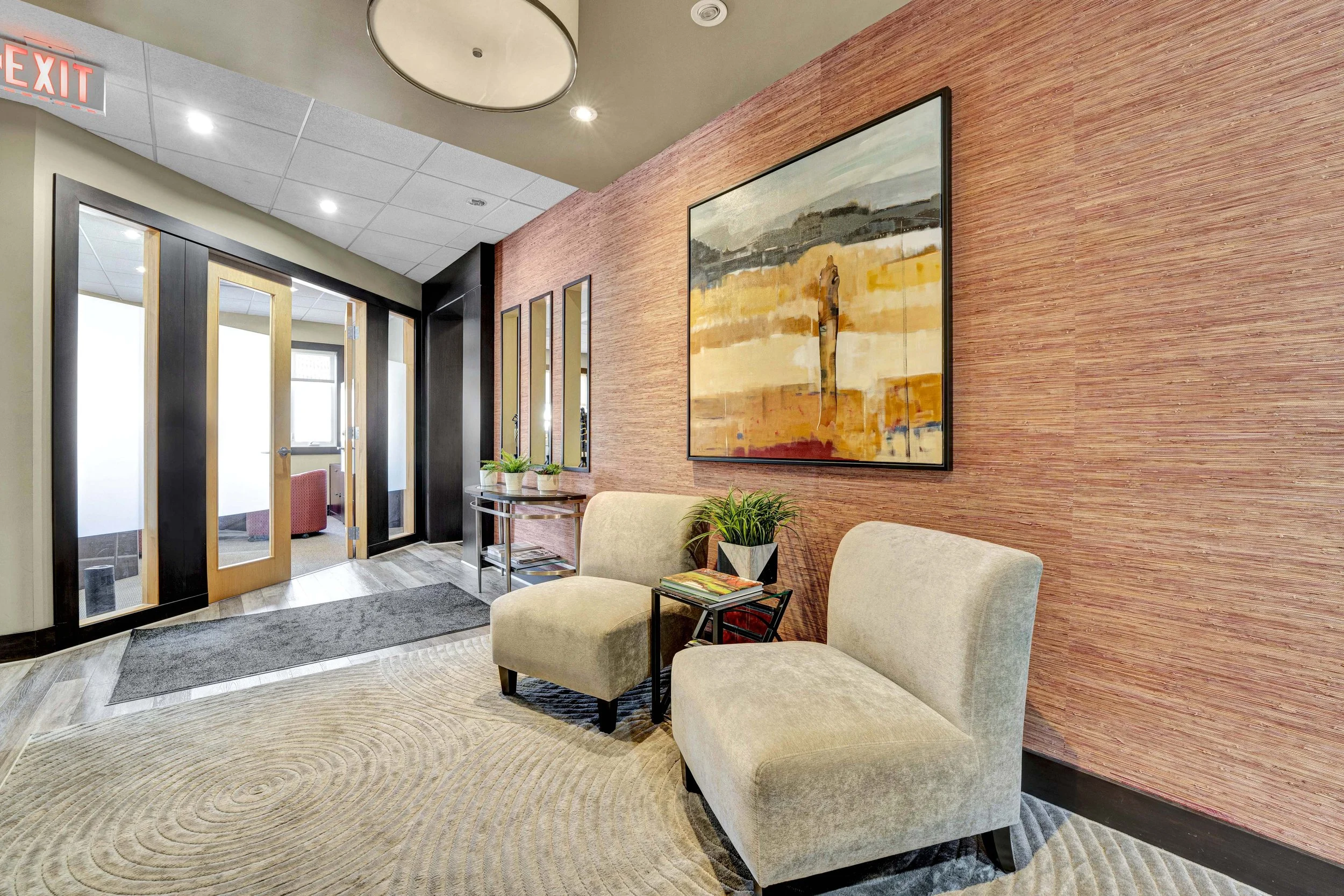 Ottawa Real Estate Photographer NVISA waiting area with beige armchairs, a side table with magazines, a large abstract painting on the textured reddish wall, potted plants, and glass double doors leading to another room, in a modern office or clinic.