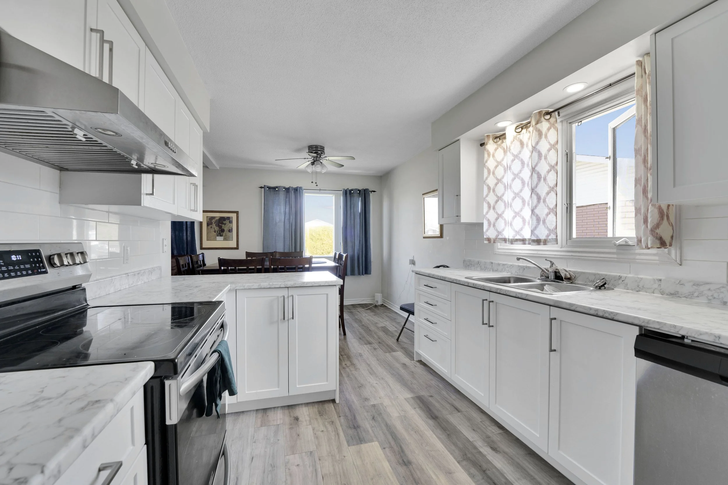 Ottawa Real Estate Media Services  A bright kitchen with white cabinets, marble countertops, a stainless steel stove, and large windows with curtains. There is a dining area with chairs visible in the background.