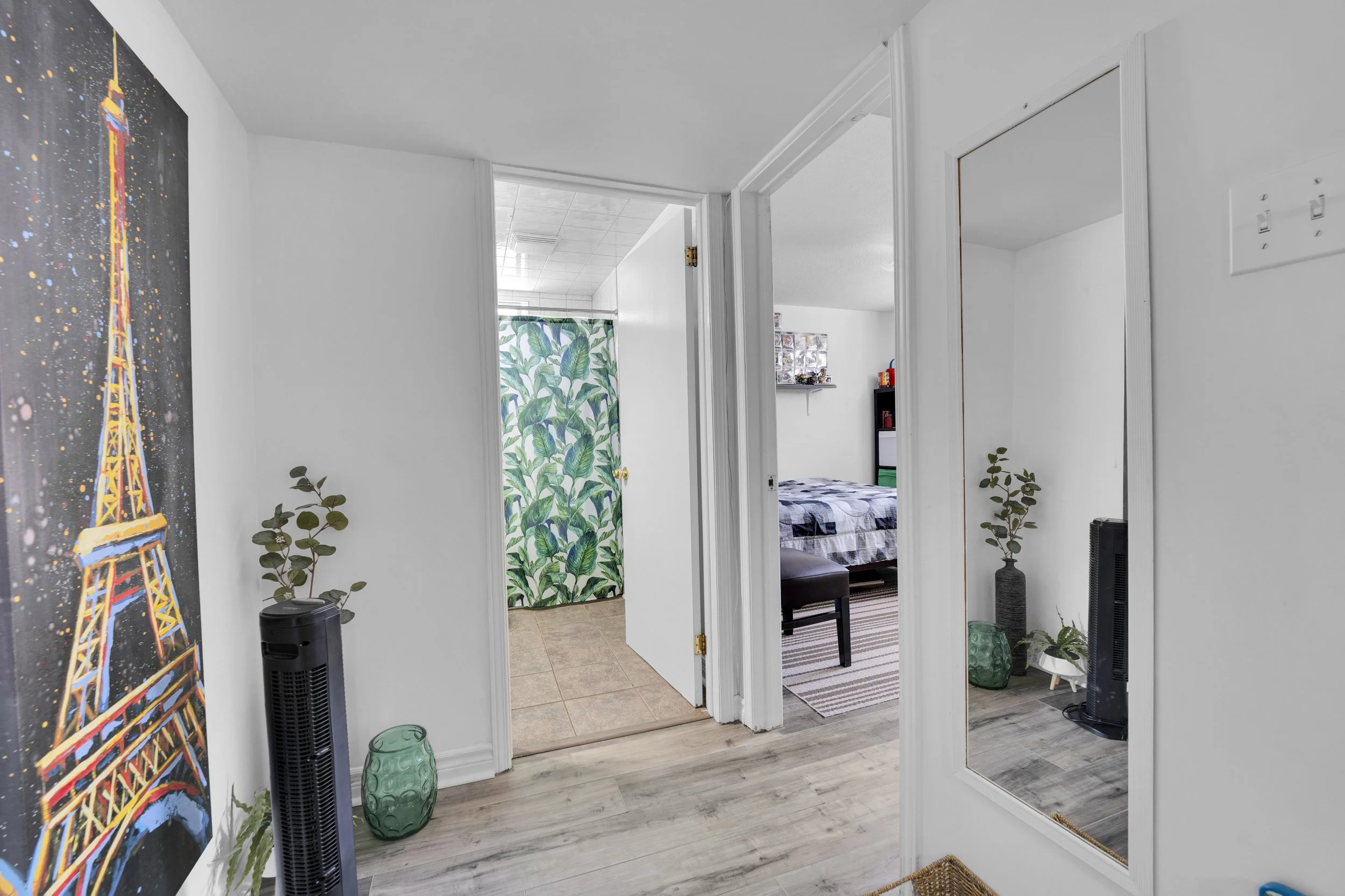 A hallway in a home with a colorful painting of the Eiffel Tower on the left wall, a full-length mirror on the right, and a view into a bedroom with a bed and a small table. There are two green vases and potted plants on the floor, and a black tower 
