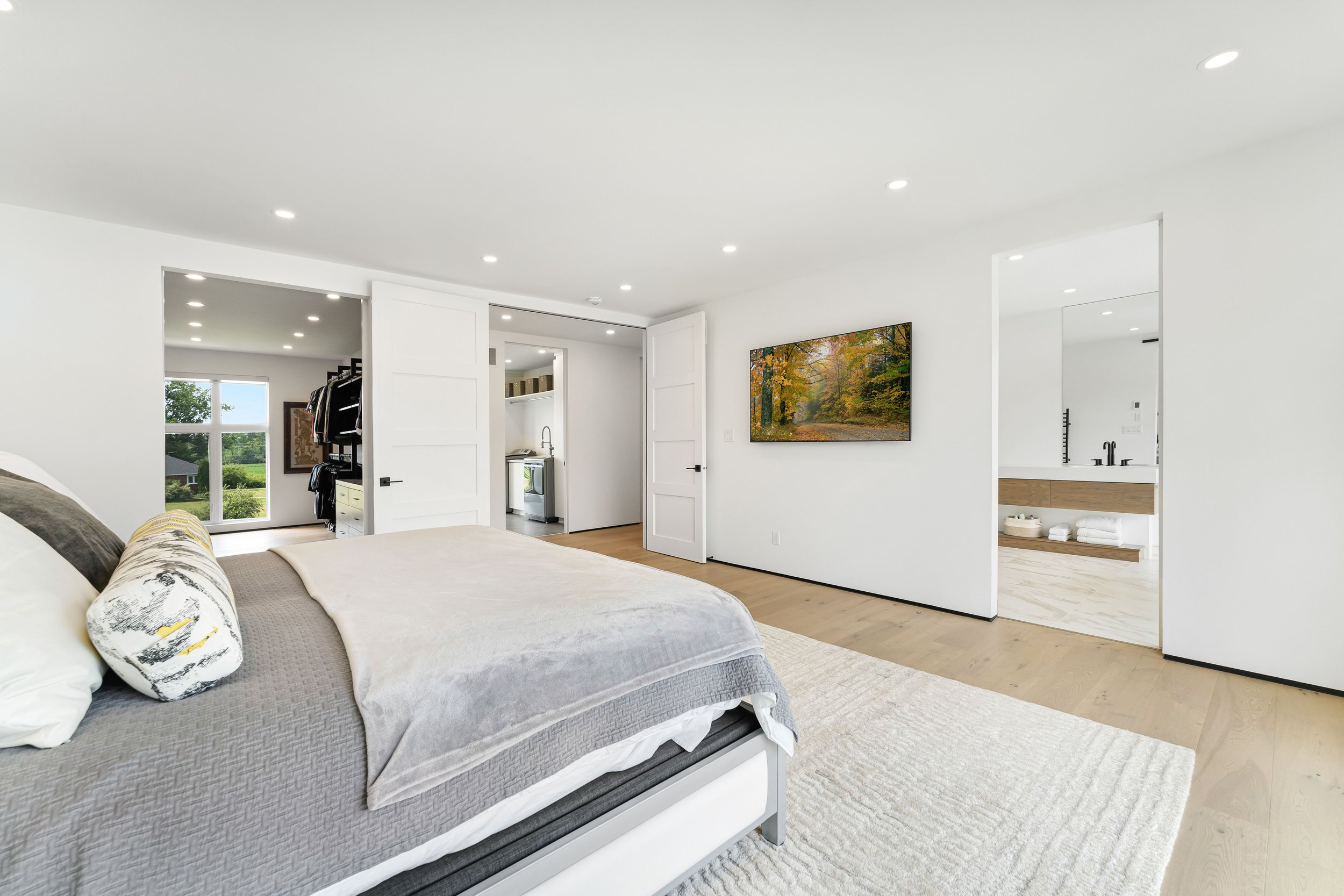 Modern bedroom with a large bed, neutral tone bedding, and a plush white rug on light wooden flooring. The room includes a walk-in closet, a wall-mounted landscape painting, and a view into a bathroom. Recessed lighting and a large window provide amp